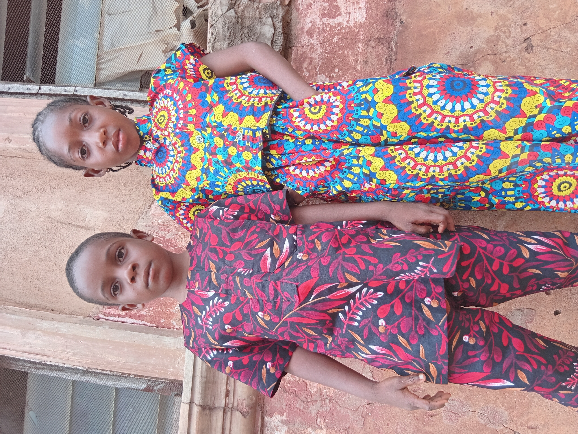 Two children are captured in a vertical, full-body portrait, standing outdoors against a weathered reddish-brown wall in what appears to be Shagamu, Nigeria. Both children, of African descent, are looking directly at the camera with neutral expressions.
The child on the left, taller and seemingly older, is wearing a vibrant, short-sleeved dress featuring a striking pattern of concentric circles and swirling mandala-like motifs in bright blue, yellow, and red. The dress has a collar and buttons running down the front. This child's hair is styled in small, dark braids.
The child on the right, shorter and seemingly younger, is dressed in a matching two-piece ensemble consisting of a short-sleeved top and full-length pants. The fabric is adorned with a dense, leafy, or floral pattern in deep red, black, and accents of orange and white. This child has short, dark hair.
The background includes parts of a window with a metal mesh grill visible behind the taller child. The wall itself shows signs of wear, with chipped paint or plaster. The ground beneath them is a light reddish-brown, possibly dirt or unpaved surface. The natural lighting indicates it is daytime. There is no visible text in the image.