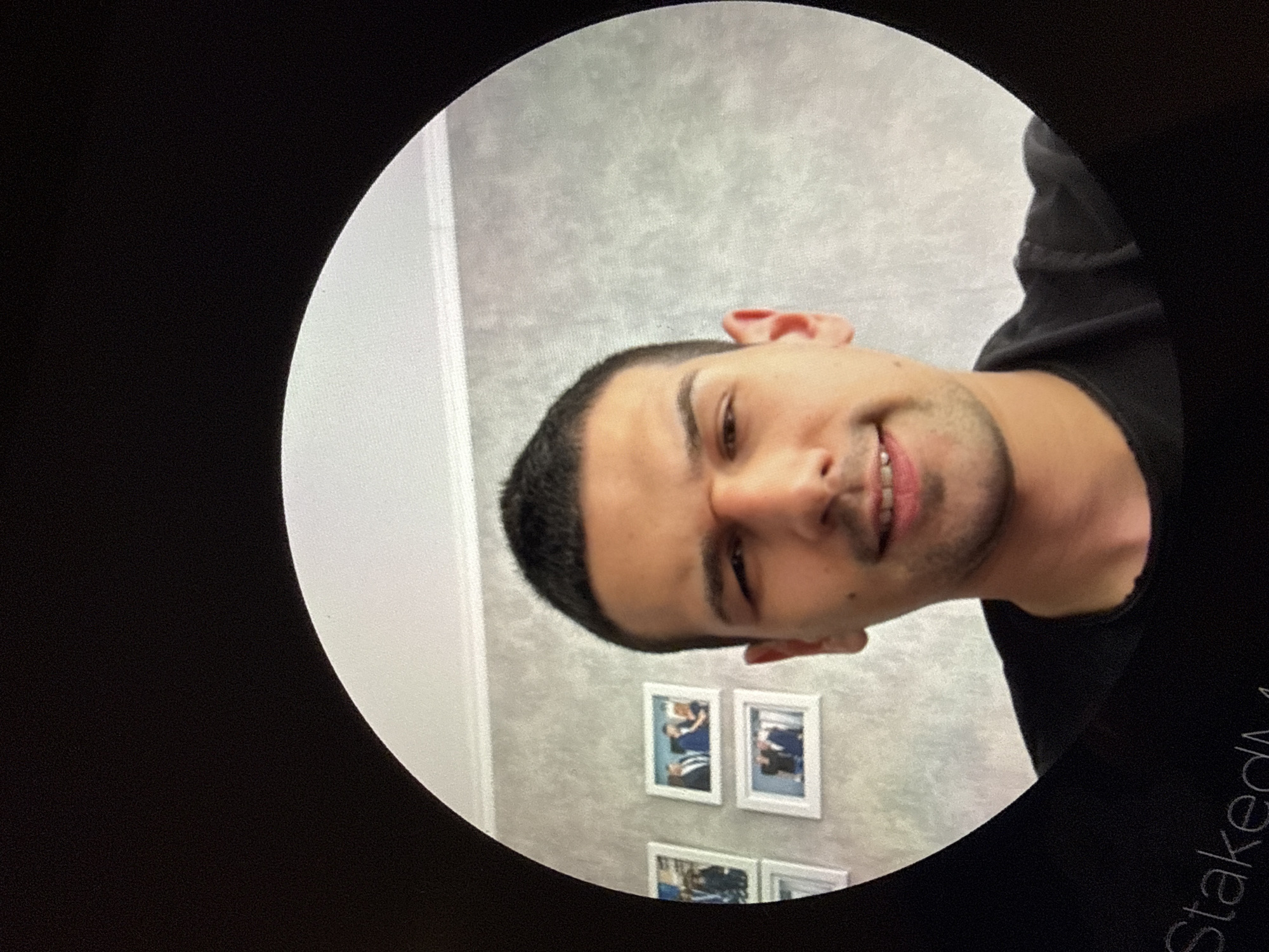 A close-up, indoor portrait shot of a man, viewed through a circular frame, possibly a camera lens or effect. The man has short, dark hair and light skin. He's wearing a black shirt. He is smiling slightly, facing the camera. Behind him is a light grey wall. Three framed pictures are visible on the wall behind him. The picture appears to be taken indoors and a portion of the word “Staked” is visible at the bottom. The city of Lovech, Bulgaria cannot be verified as a location.