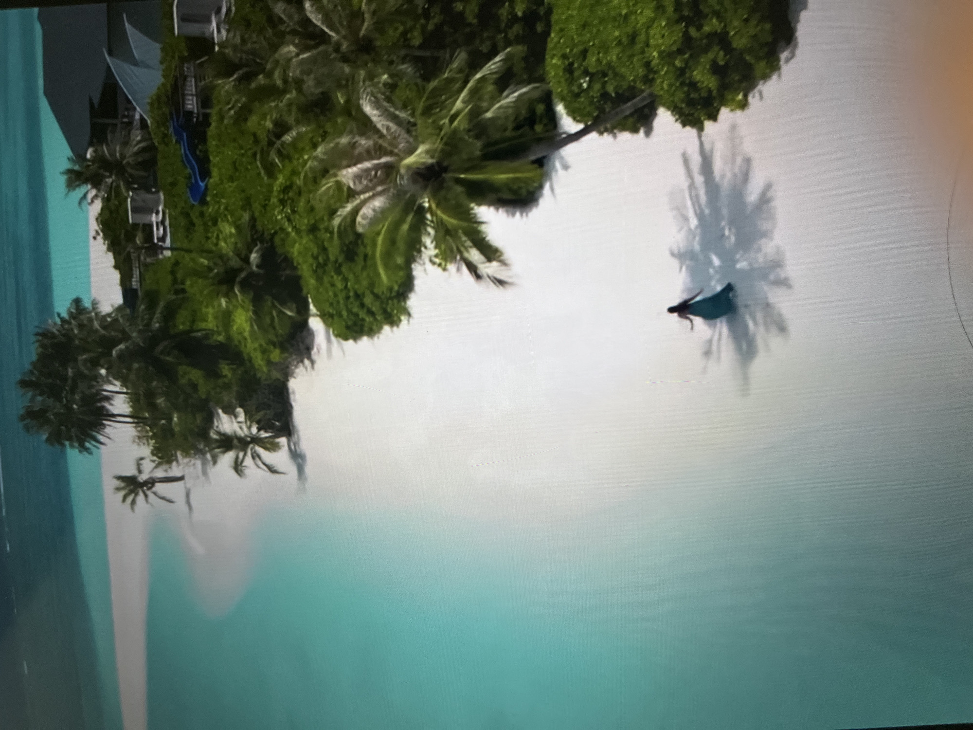 An aerial view depicts a pristine white sand beach meeting turquoise ocean waters. A lone figure, dressed in a flowing teal gown, walks along the shoreline. Lush green vegetation, including palm trees, borders the beach, with a modern resort structure and a blue waterslide visible among the foliage. The scene suggests a tropical paradise.