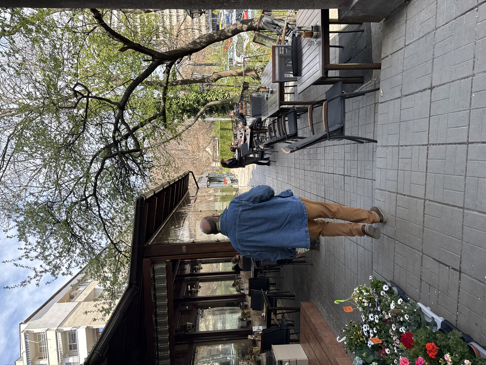 The scene captures an outdoor dining area along a sidewalk in Sofia, Bulgaria. The area is partially covered by an awning extending from a building, likely a restaurant or café. A man with a bald head, wearing a blue denim jacket and brown pants, walks along the sidewalk. There are tables and chairs set up for outdoor dining, with some patrons seated at the tables. Trees with budding leaves provide shade and add to the outdoor atmosphere. Flowers are in a bucket on the sidewalk. The ground is paved with grey brick tiles.