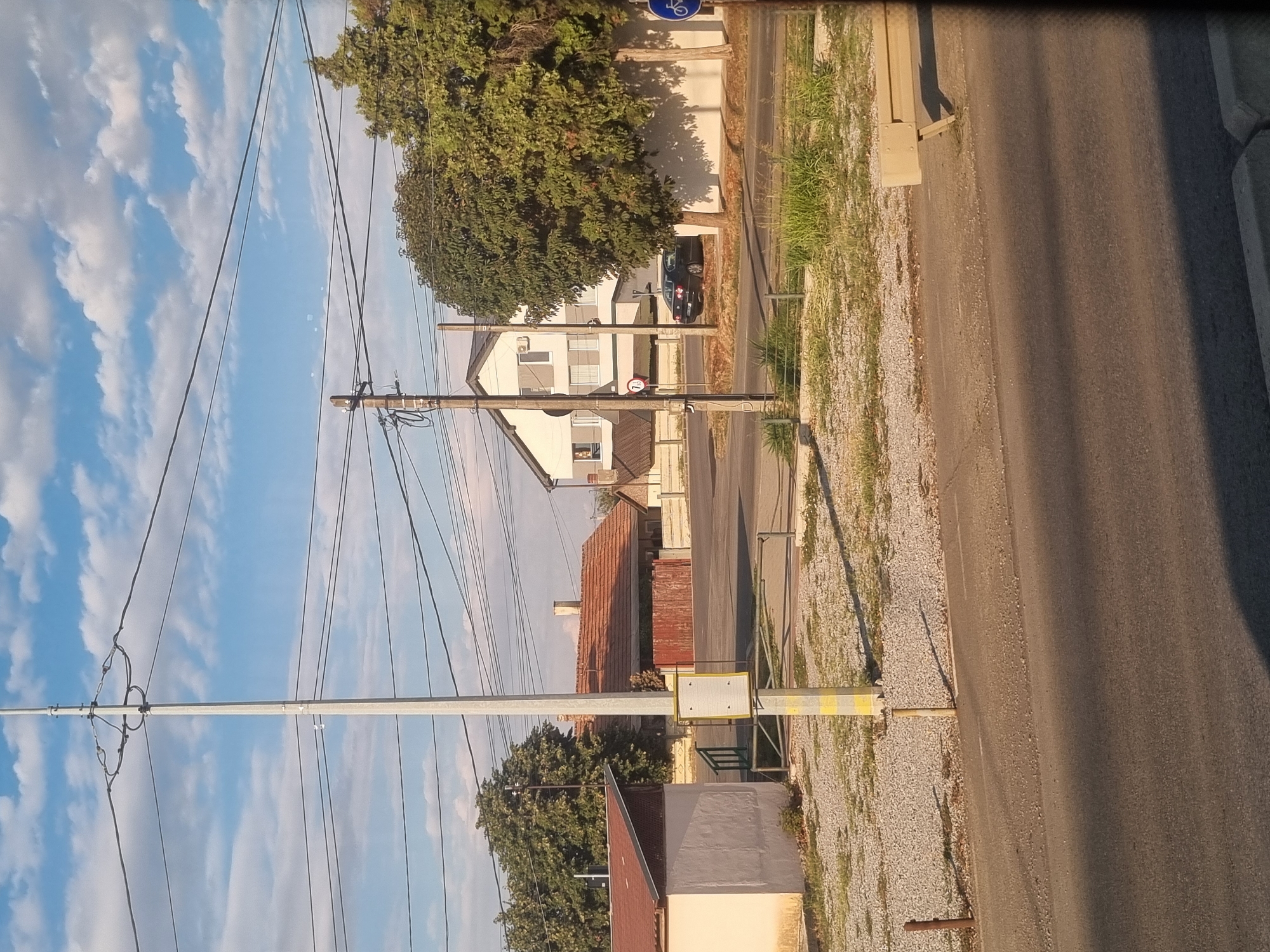 The image depicts a street scene in Sântion, Romania, under a partly cloudy sky. A tall utility pole with numerous wires dominates the foreground, running horizontally across the frame. To the left, a low, light-colored building with a brown roof is partially visible. Behind it, a house with a terracotta tiled roof and a wooden fence is situated. Further back, a modern, multi-story white house with dark windows stands. A dark-colored sedan is parked on the right side of the street in front of a white wall and a large, leafy tree. The street is paved, with a gravel-covered area to the left featuring some sparse vegetation. A bench is visible on the right side of the pavement, and a blue sign with a white bicycle icon is also present. The lighting suggests it is late afternoon, with long shadows cast across the scene. There are no people visible in the image.