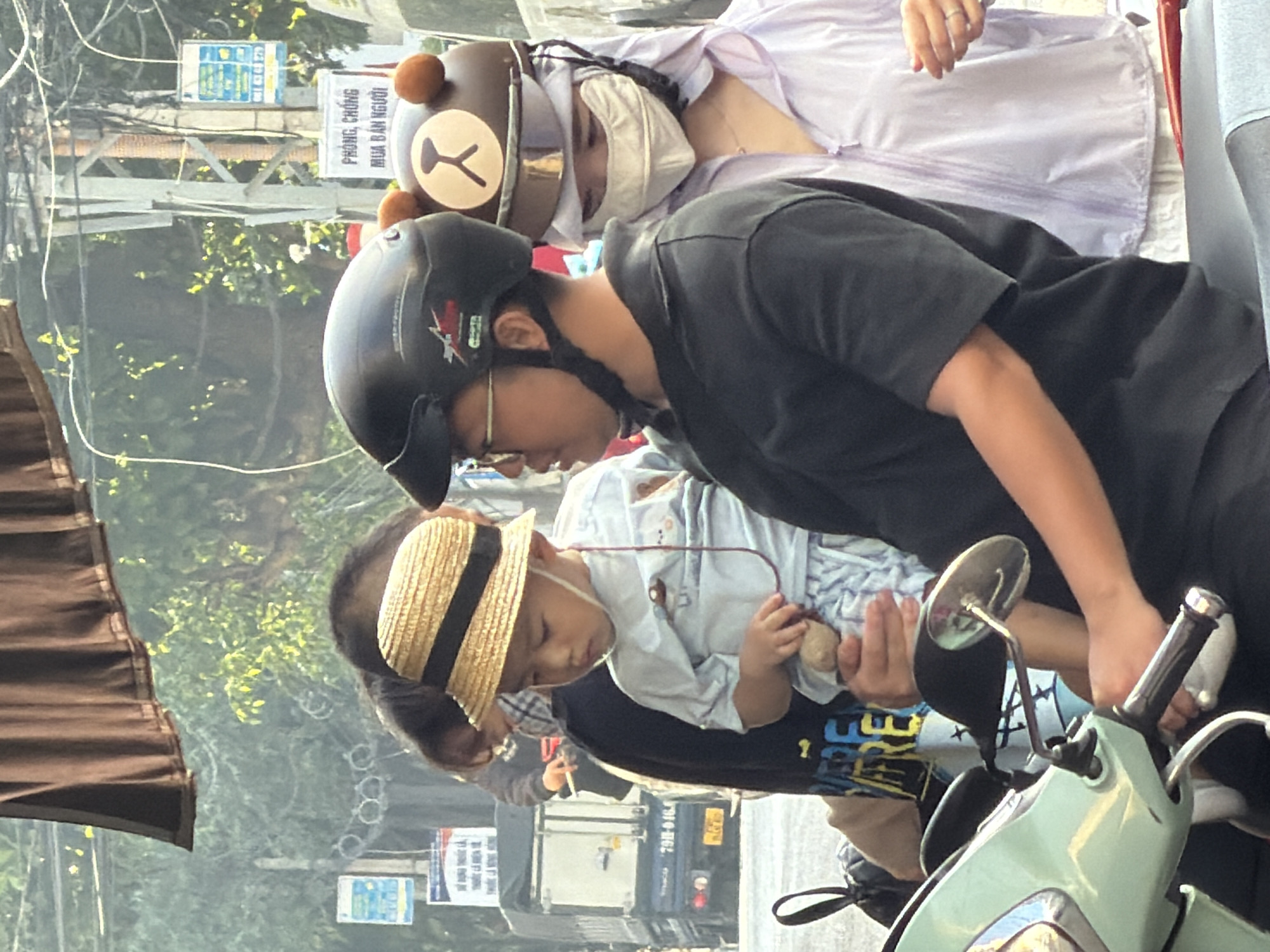 The image depicts a street scene in Nha Trang, Vietnam, where a group of people are riding a scooter. The scooter has a light green body and a black seat. In the foreground, a person wearing a black shirt and a black helmet is driving the scooter. They are holding a small child in their arms who is wearing a straw hat and a light blue outfit. To the right of the driver, another person is riding on the back, wearing a light purple jacket and a white mask covering their face. This person also has a helmet adorned with a bear face design. In the background, there are vehicles, signs, and trees, suggesting an urban environment. The lighting and atmosphere indicate it is daytime.