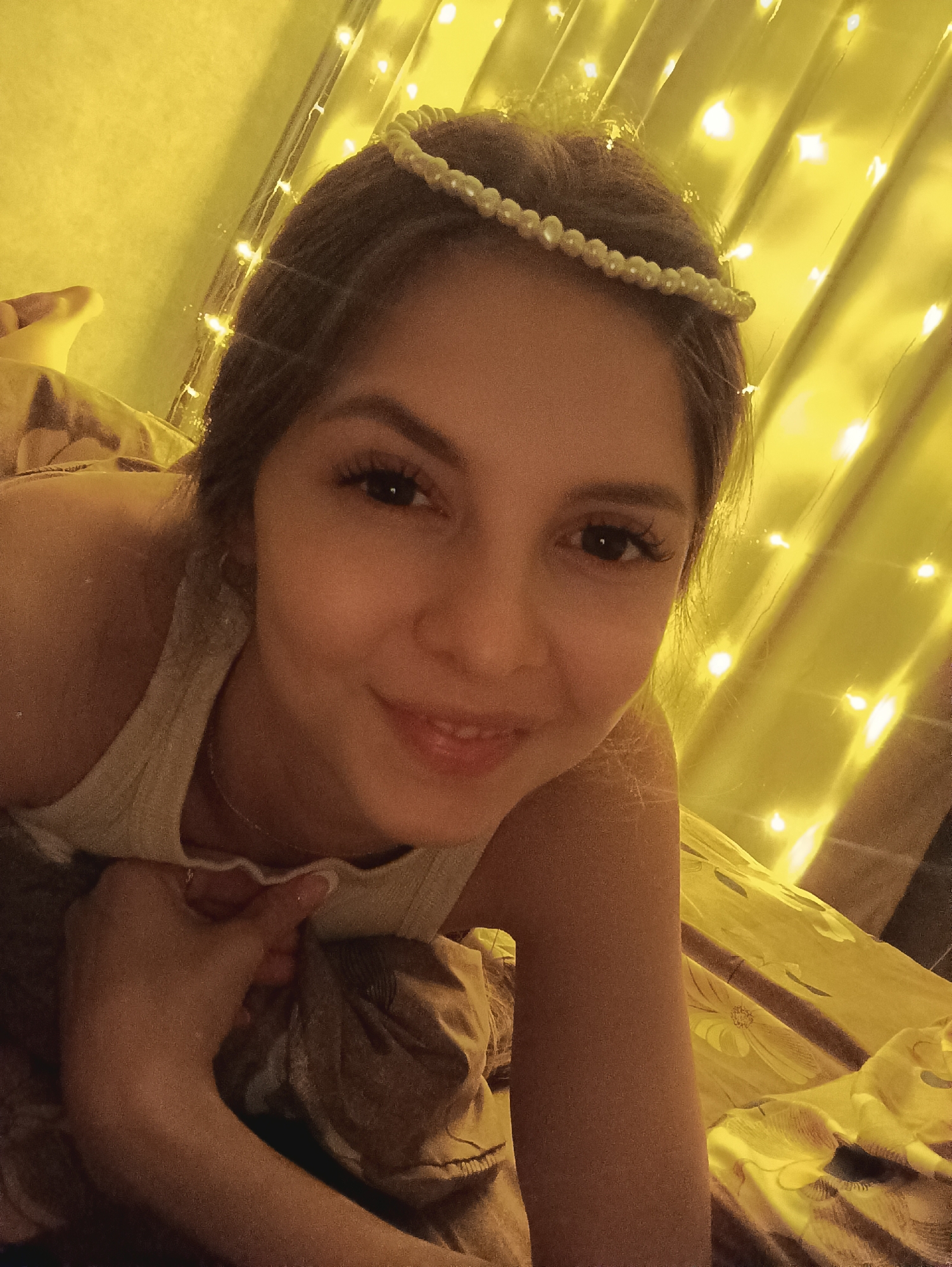 A person is lying on a bed. The person has dark hair and is wearing a pearl headpiece. The person is smiling and looking at the camera. The bed is covered in a brown floral print. There are string lights behind the person. The location is Unknown, Unknown.