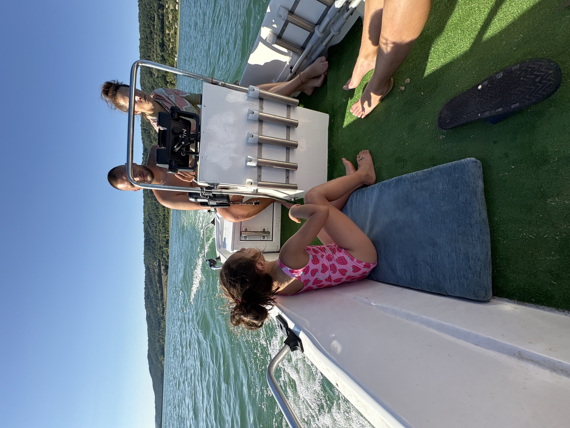 An outdoor, mid-angle, eye-level shot captures a family enjoying a boat ride on a sunny day in the city of Golyama Zhelyazna, Bulgaria. The boat is moving through a body of water with green, choppy waves, and a forested shoreline with hills rises in the background under a clear blue sky. In the foreground, a young girl with dark hair pulled back in a ponytail sits facing the stern of the boat, wearing a pink swimsuit adorned with strawberries. She is seated on a dark blue cushion on the green carpeted floor of the boat. To her right, two adults, a man and a woman, are seated towards the front of the boat, facing forward. The man, with short hair and a beard, is partially obscured by the woman. The woman, with her hair tied up, is looking towards the boat's console, which has a Garmin GPS device. Their legs are visible, and the woman is wearing a light-colored, patterned top. Other individuals' legs are also visible in the foreground on the right, indicating at least two more people are on the boat. A black sandal lies on the green carpet near the right edge of the frame.