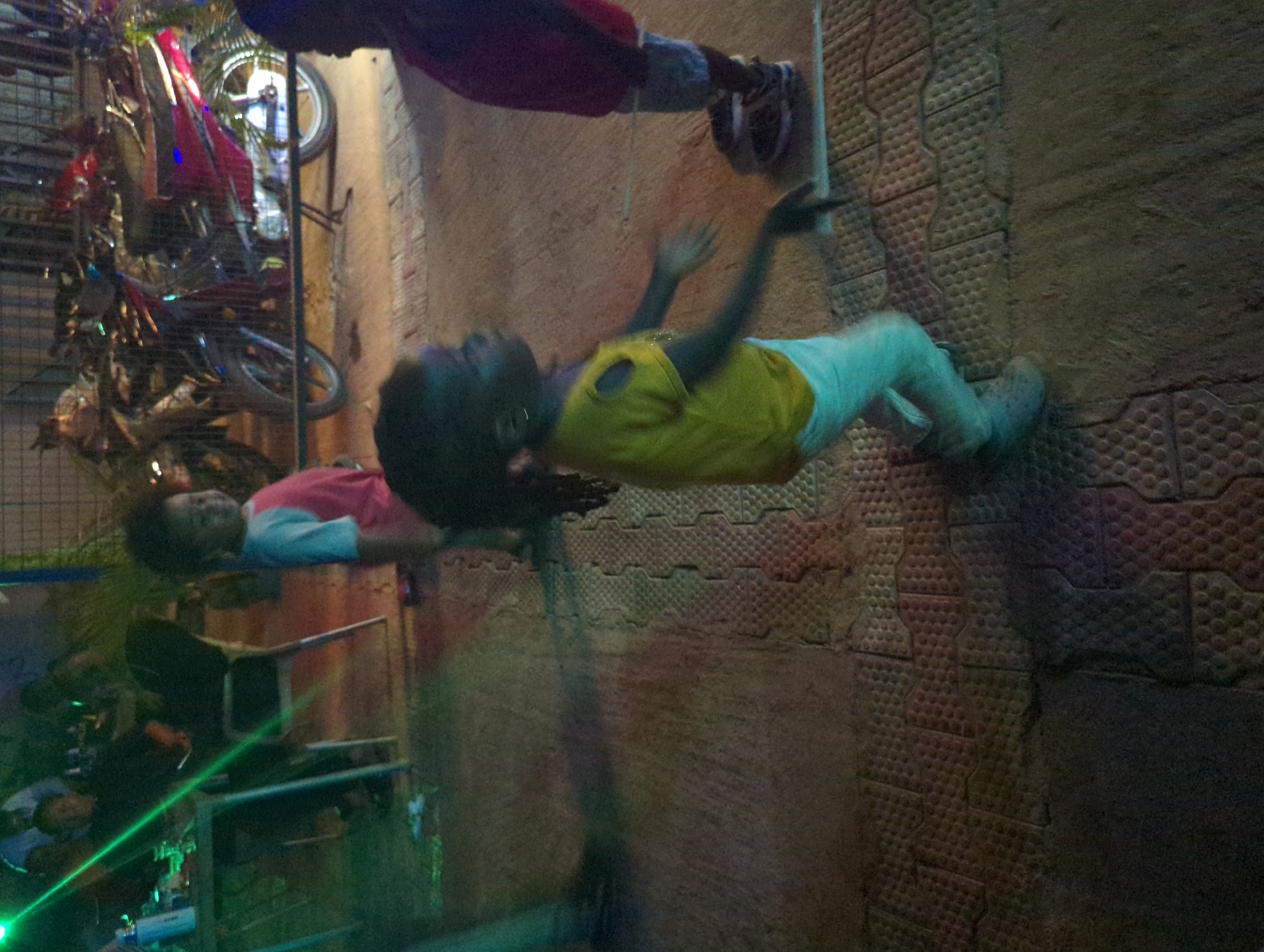 The image shows two children on a textured surface. One child is lying down, wearing a pink and blue shirt. The other child is standing, wearing a yellow shirt and light-colored pants, with arms raised. A third person is partially visible above the standing child, wearing dark clothing. The scene appears to be outdoors on a paved surface with a distinctive pattern of raised circles. Behind the children, there are motorcycles parked behind a metal fence and a table with what looks like laser lights and bottles. Given the context, this is likely a residential area in Narayi, Nigeria.
