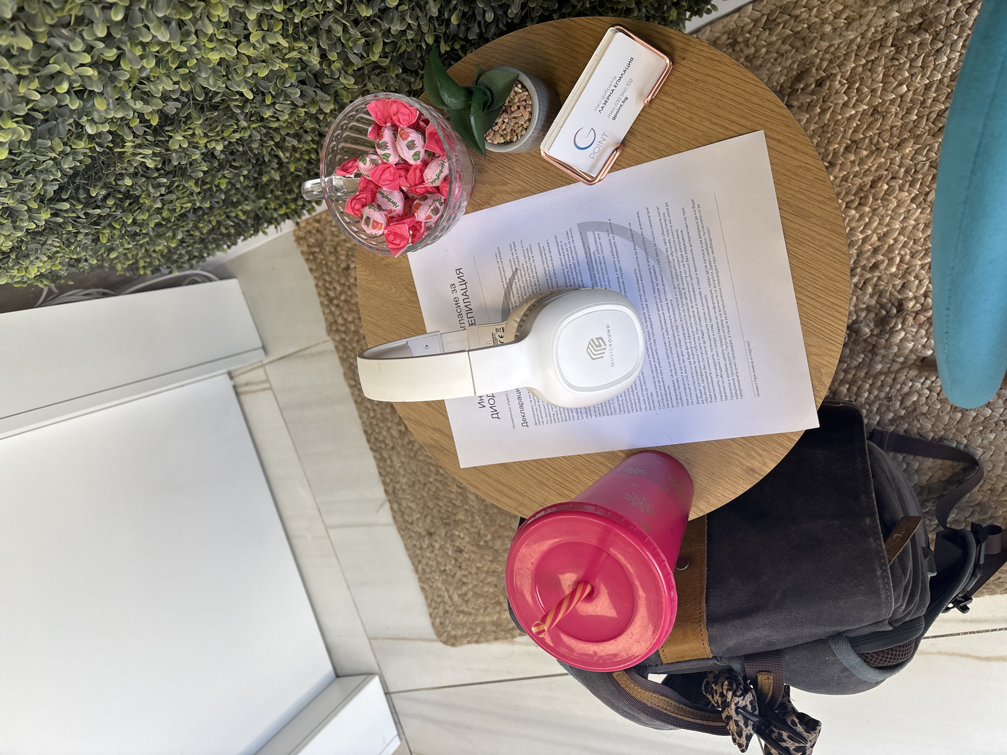 A high-angle, indoor shot captures a round, wooden table displaying an assortment of items. On the left, a glass bowl filled with pink, strawberry-flavored Herbafielda candies sits beside a small potted plant. A white business card for "G POINT - ЛАЗЕРНА ЕПИЛАЦИЯ," located in Plovdiv, Bulgaria, is partially visible. Below these items lies a document titled "Декларация за ЕПИЛАЦИЯ" (Declaration for Epilation), with the words "MUSICSOUND" visible on a pair of white headphones. Further down, a pink tumbler with a straw is placed next to a gray backpack with brown leather accents and a leopard-print accessory. The table is positioned on a patterned rug, adjacent to a blue chair. In the background, a white wall and a green wall decorated with artificial plants are visible.