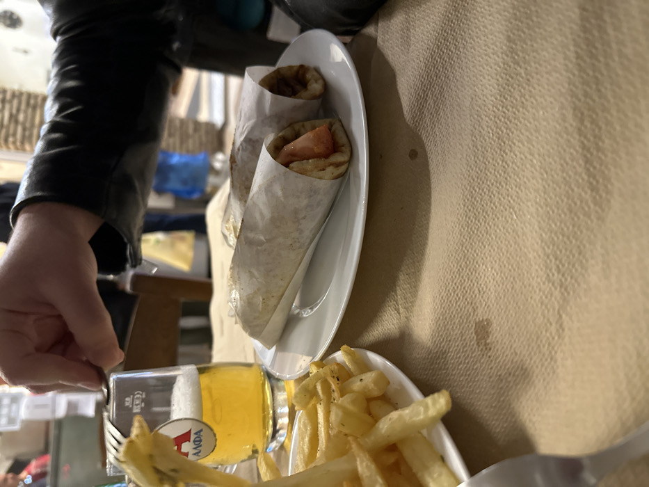 The image presents an indoor dining scene from a slightly tilted, overhead perspective, likely in a casual restaurant or eatery in Nea Erithrea, Greece.
On the left, a person is partially visible, wearing a black leather jacket. Their left hand holds a metallic fork, poised above a glass of beer, indicating they are about to eat or are in the process of eating.
The table is covered with a light brown, textured tablecloth. The meal consists of classic Greek fare. Two gyros or souvlakis, wrapped in paper, are served on a white oval plate. One of the wraps clearly shows red tomato slices as part of its filling. Next to these, a separate small white dish is filled with golden french fries, which appear to be seasoned with herbs or spices.
A glass of light-colored beer is prominently featured, with a white and red label clearly displaying the Greek word "ΑΛΦΑ" (Alpha), a recognizable Greek beer brand, confirming the local setting.
The background is softly blurred, showing vague hints of the surrounding restaurant interior, including what might be furniture or other patrons, contributing to a casual dining atmosphere. The overall scene captures a moment during a meal, emphasizing typical Greek comfort food.