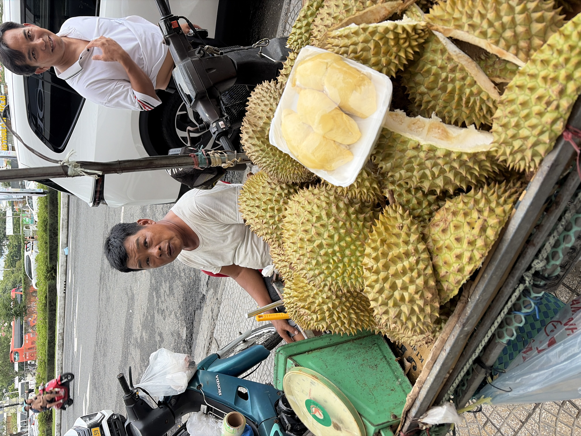 A middle-aged East Asian man with short black hair and a light-colored t-shirt sits on a red stool next to a cart filled with durians. To his right, another East Asian man wearing a white polo shirt stands and adjusts his shirt. The cart also contains a green weighing scale and a white plastic container with durian flesh. A motorcycle is parked to the left of the man in the t-shirt. The scene appears to be on a city street in Đà Nẵng, Vietnam, with a white SUV and other vehicles in the background. The ground is paved with light-colored tiles.