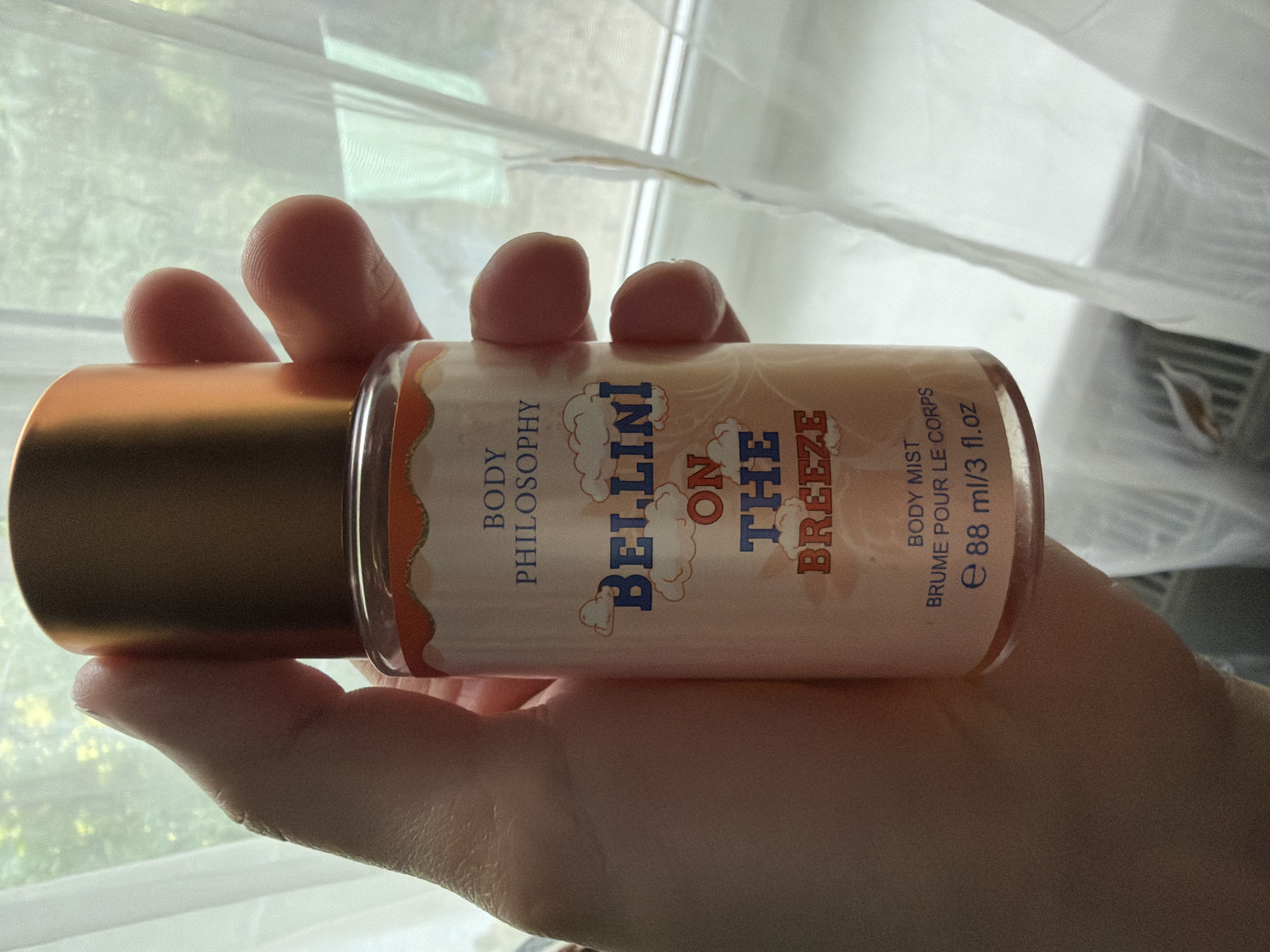 A hand holds a bottle of Philosophy Bellini On The Breeze body mist. The bottle is made of clear glass with a peach-colored label featuring orange cloud-like accents and blue text. The text on the label reads "BODY PHILOSOPHY", "BELLINI ON THE BREEZE", and "BODY MIST BRUME POUR LE CORPS e 88 ml/3 fl.oz". The bottle has a metallic gold cap. The background is blurred and appears to be a window with sheer white curtains.