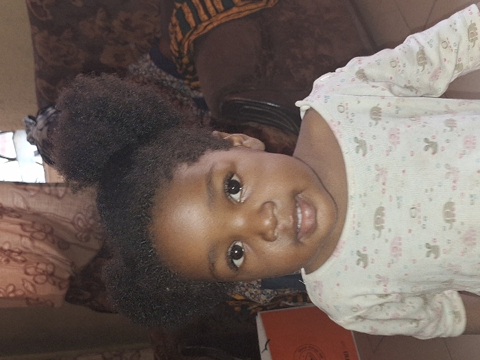 A young child, approximately 1-3 years old, with dark skin and dark, Afro-textured hair styled into two prominent puffs, faces forward. The child wears a light-colored, long-sleeved top patterned with small pink elephants and other indistinct shapes. The child's gaze is directed towards the viewer with a neutral expression and slightly parted lips. The setting is an indoor residential space in Kaduna South, Nigeria. In the background to the right, a portion of a brown upholstered sofa or armchair is visible, draped with a patterned cloth featuring black, orange, and yellow geometric designs. A translucent brown curtain with a floral pattern partially covers a window in the upper right background. To the left, a segment of an orange bag or box with white lettering is visible. The floor is covered with light-colored tiles. Ambient lighting illuminates the scene.