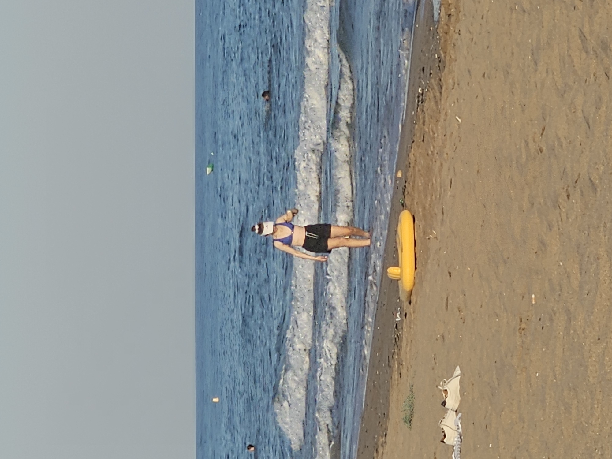 A woman wearing a blue bikini top, black shorts, and a white visor walks in the shallow water near the shore of a beach. She is holding a white object in her right hand. Two sets of white sneakers are on the sand in the foreground, to the left. A yellow inflatable ring is partially visible on the sand to the right of the woman. The sea is blue with white waves, and the sky is clear and pale blue. Two buoys are visible in the water, one yellow and one green and white. Another person is partially submerged in the water further out in the sea. The scene appears to be at Platja de Castelldefels, Spain.
