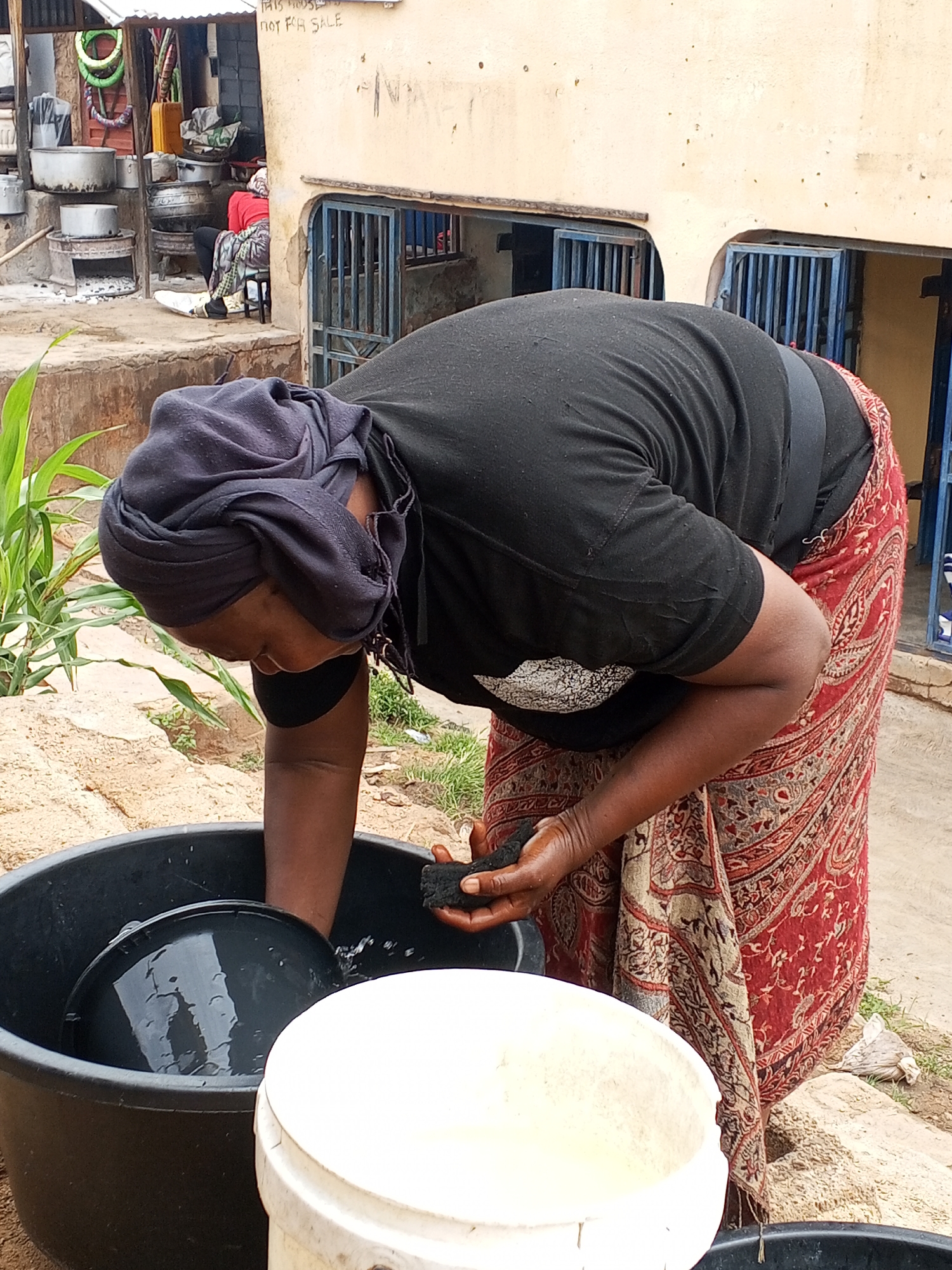 In Gwong, Nigeria, a woman is outside washing a black plastic tub filled with black pots. She wears a black shirt, a patterned red and beige skirt, and a dark head covering. She's holding a dark sponge or cloth in her right hand, presumably for cleaning. Next to the black tub is a white bucket. In the background is a person sitting next to large cooking pots near an open fire. The environment is a yard or outdoor space next to a building with metal bars on the windows. A sign above the windows is partially visible, with the words "NOT FOR SALE."