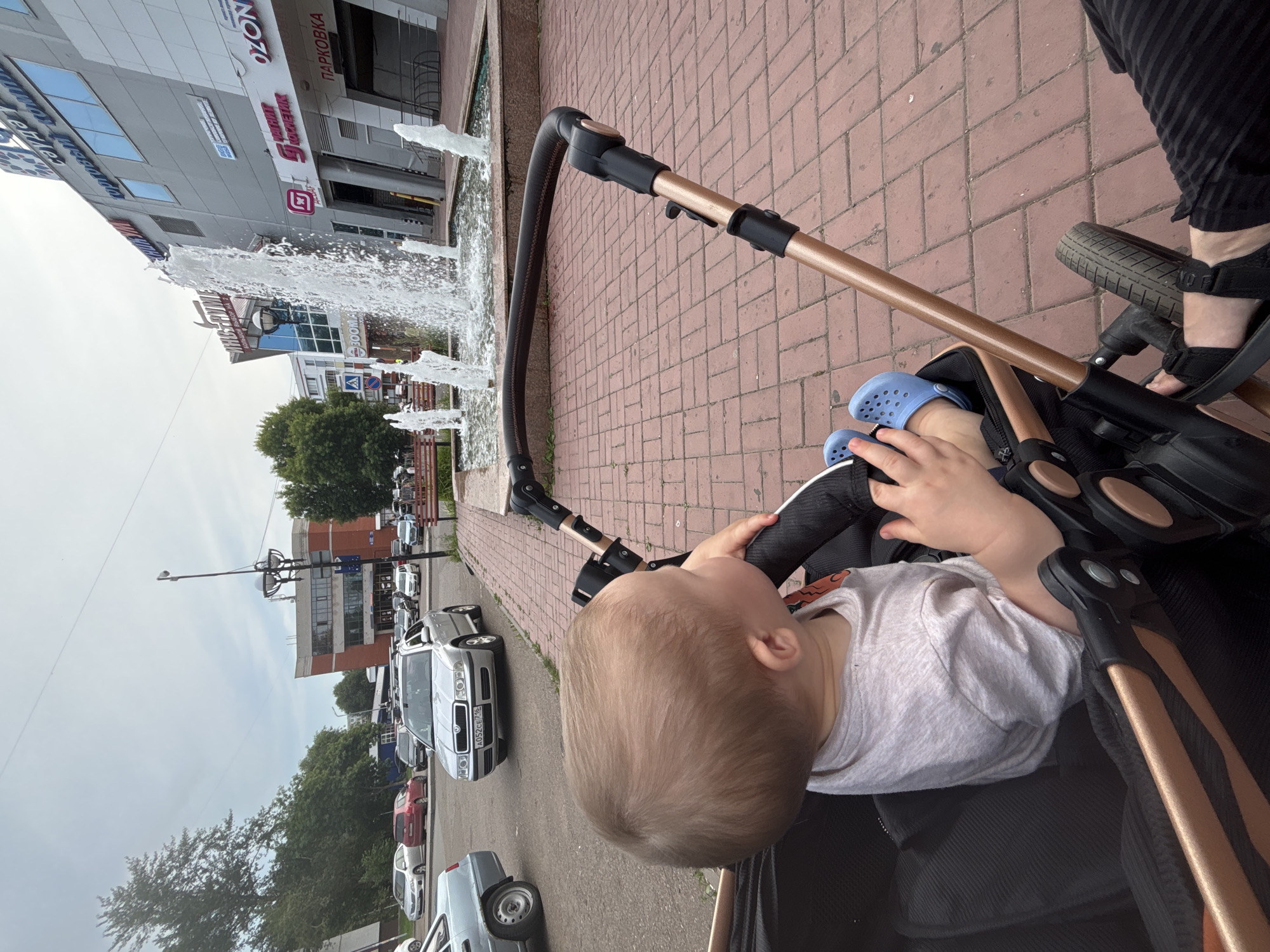 A young child sits in a stroller on a brick-paved sidewalk in front of a modern building with a large fountain. The child has light brown hair and is wearing a light-colored t-shirt and blue Crocs. The stroller is black with gold accents. In the background, a silver car is parked on the street, and trees line the sidewalk. The building has multiple stories and appears to be a commercial establishment with several signs, including "OZON" and "ПАРКОВКА" (Parking). The sky is overcast. The location is identified as Naberezhnye Chelny, Russia.