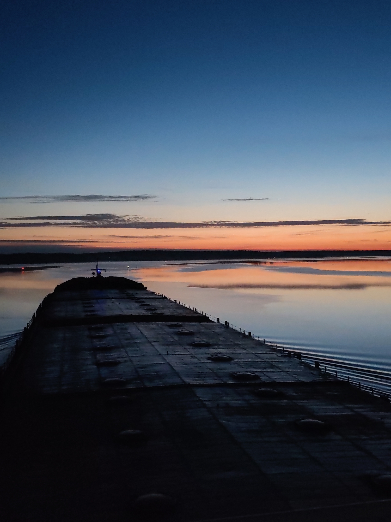 The image depicts a twilight scene from the deck of a large cargo ship. The deck is dark and appears to be made of metal plates. In the distance, a body of water reflects the orange and pink hues of the setting sun. A dark shoreline is visible in the background. The sky transitions from a deep blue at the top to orange and pink near the horizon. There are a few thin clouds scattered across the sky. The location is not identified as Nova, Russia. There is no indication of people in the frame.