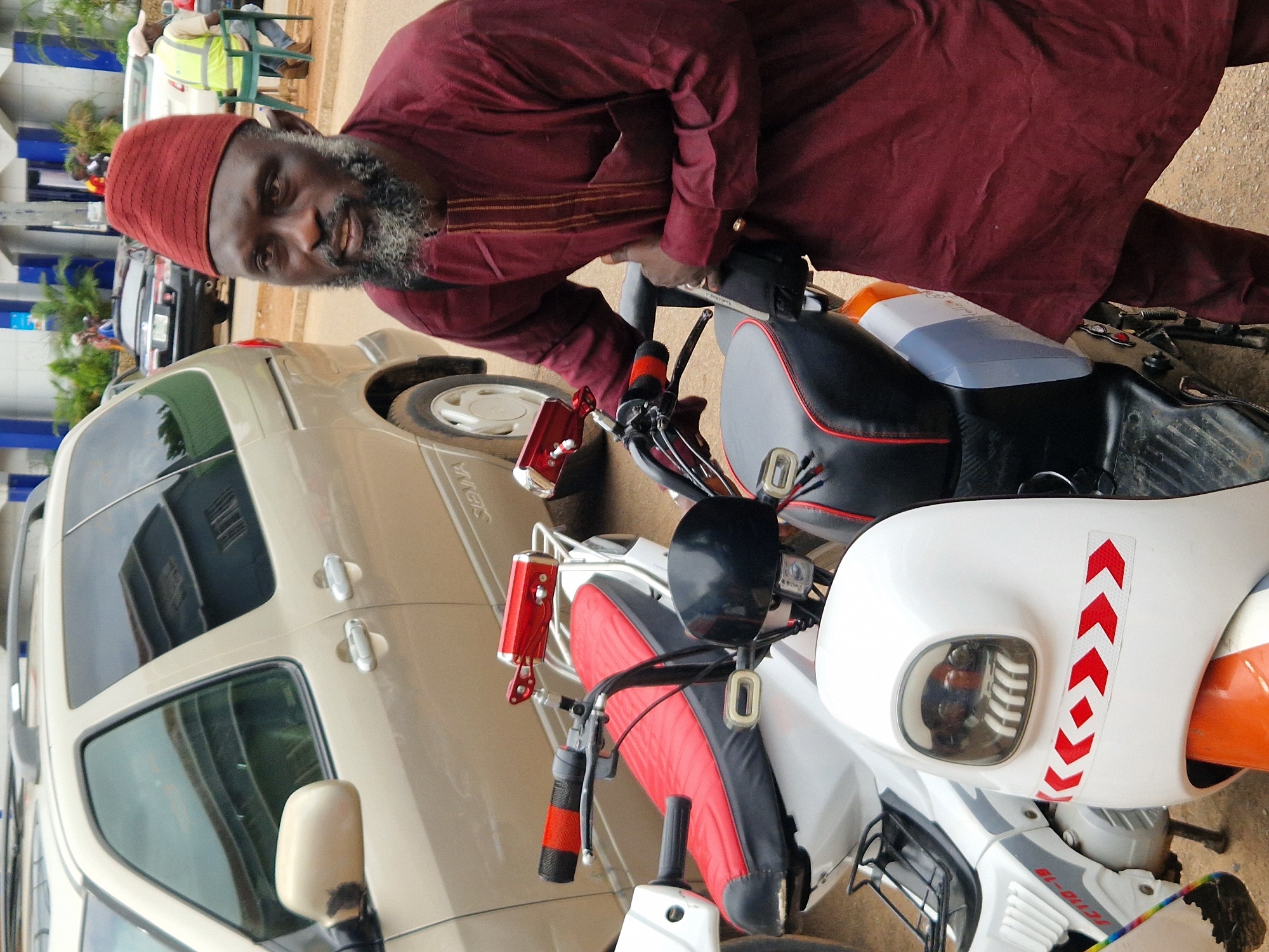 A medium shot captures a man with a salt-and-pepper beard and a red kufi hat standing next to two parked scooters and a tan minivan. The man wears a dark red kaftan, and his right hand rests on the handlebars of the scooter closest to him, while his left hand is on his stomach. The scooter features a red and black seat, a red rearview mirror, and a white front fairing with red chevron stickers. The minivan, a Toyota Sienna, is visible in the background to the left. The ground is a dirt road. In the far background, a person in a high-visibility yellow vest is seated on a green chair.