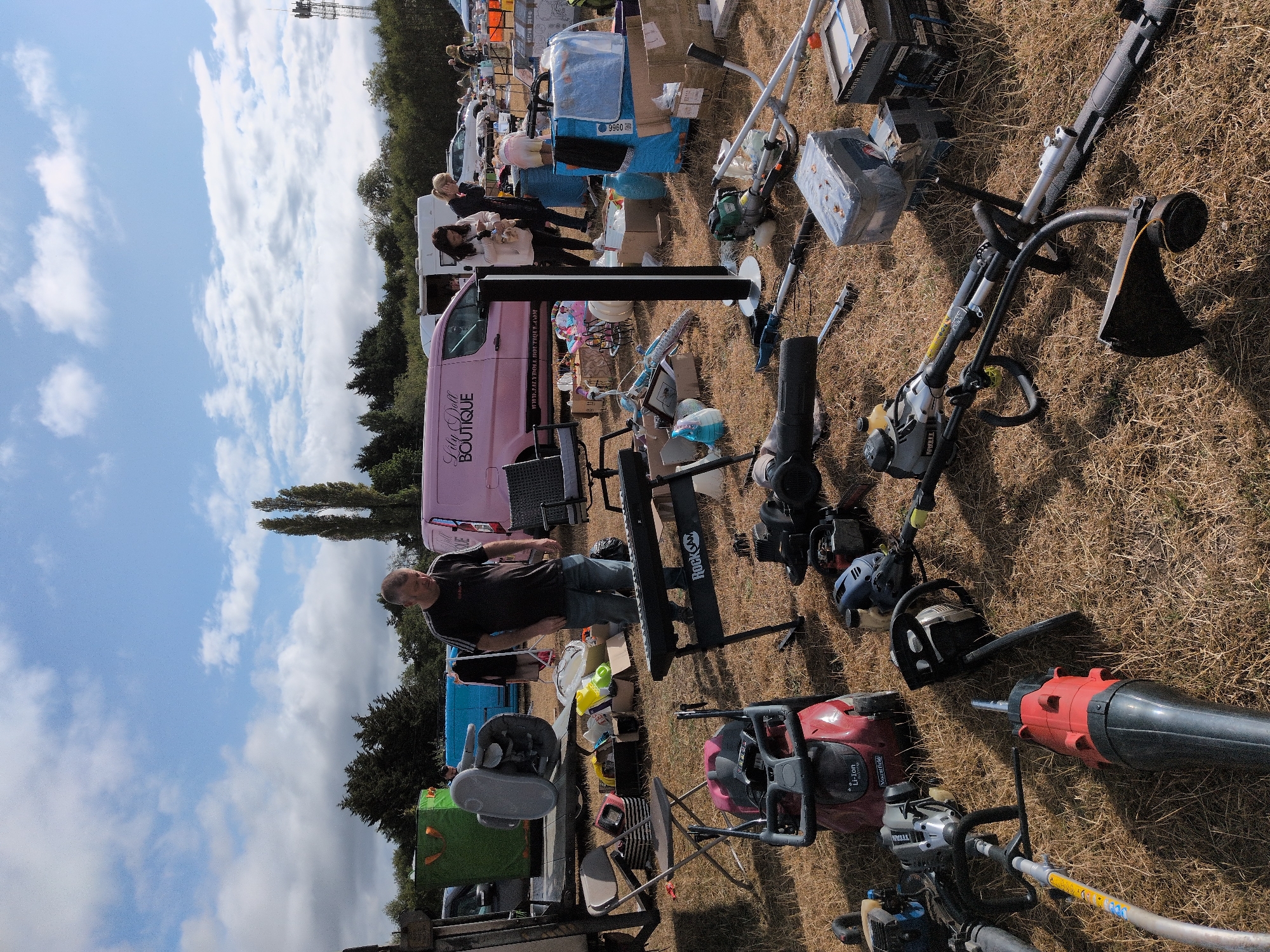 This image depicts an outdoor market, likely a car boot sale, on a dry, grassy field under a partly cloudy sky. The scene is situated in South Staffordshire, United Kingdom. A variety of items are displayed for sale, including gardening equipment such as lawnmowers, strimmers, and leaf blowers, as well as other miscellaneous objects like a keyboard, chairs, boxes, and bags.
In the foreground, several pieces of gardening equipment are laid out on the grass, including a red and black lawnmower with "Li-ion" and "Mountfield" branding, and various strimmers and blowers with yellow and black accents. A black keyboard on a stand is also visible.
In the mid-ground, a man in a dark t-shirt and jeans is standing and looking down at some items. Behind him, a pink van with "Lily Doll BOUTIQUE" branding is parked. Two women are standing near the van, one holding what appears to be a small animal. A black speaker on a stand is positioned in front of the van. Various boxes and bags are scattered around the area.
In the background, more vehicles and stalls are visible, along with trees and a radio tower in the distance. The overall impression is of a bustling market day with a diverse range of goods on offer.