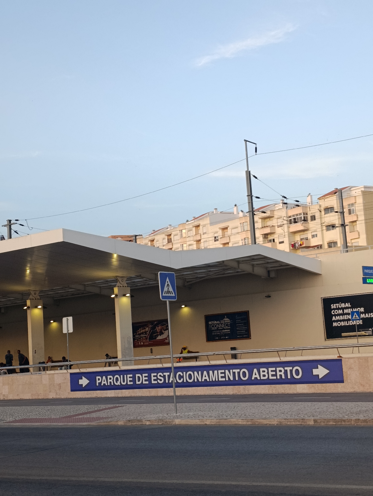 The image shows a large, open-air parking garage entrance in Setúbal, Portugal. The entrance is marked with a blue sign that reads "PARQUE DE ESTACIONAMENTO ABERTO" (Open Parking Lot) with a white arrow pointing to the right. Above the entrance, there are buildings with light-colored facades and red-tiled roofs. A pedestrian crossing sign is visible on the right side of the image, near the entrance. People can be seen sitting and standing under the covered area of the parking garage. The sky is clear and light blue.