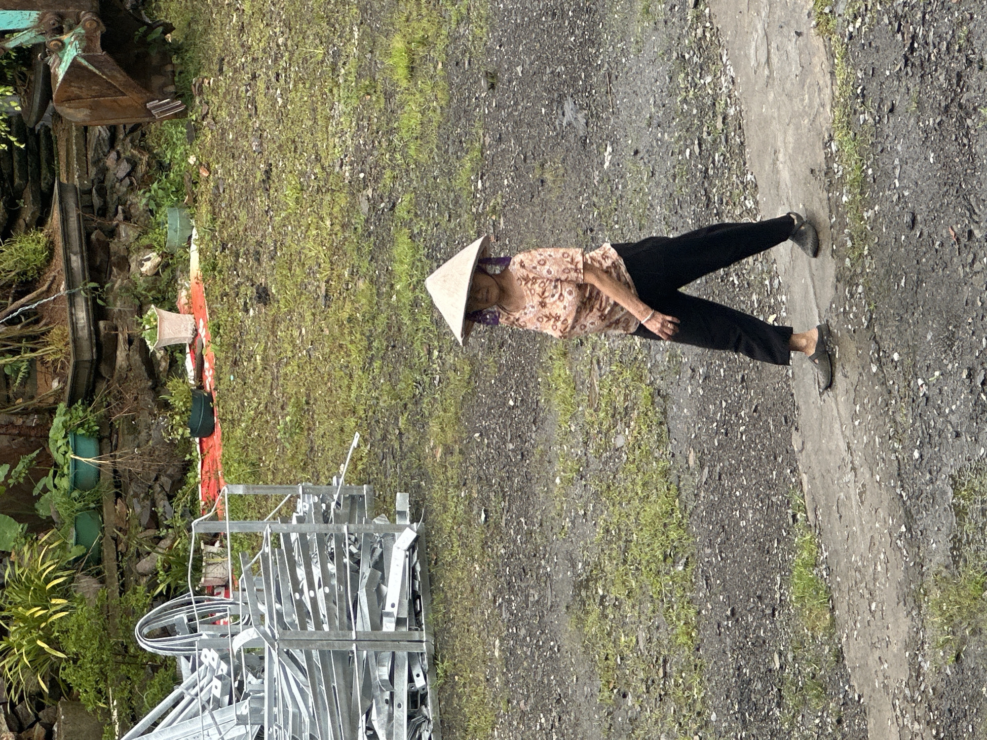 The image captures an outdoor scene, likely in Mù Cang Chải, Vietnam. The central subject is an elderly woman standing on a ground composed of dirt and gravel, interspersed with patches of green grass. She wears a traditional Vietnamese conical hat (nón lá), a light-colored shirt adorned with a dark floral or leaf pattern, dark pants, and dark flat shoes. A light-colored bracelet is visible on her left wrist.
To the woman's left, a narrow concrete path or curb runs through the frame. The background on the left side of the image reveals an area with dense greenery, including shrubs and potted plants, one of which is a light beige pot and another is a dark green pot. The rusty bucket of an excavator is partially visible in the upper left, indicating industrial or construction activity nearby. Below the excavator, a stack of silver-colored metal structures, possibly construction materials or frames, is arranged next to a red tarp.
The woman stands still, looking slightly towards her left (the viewer's left), appearing calm. The scene is brightly lit, suggesting daytime, with soft lighting indicating an overcast sky or possibly early morning/late afternoon. The setting appears to be a rural or semi-developed area. No visible text is present in the image.