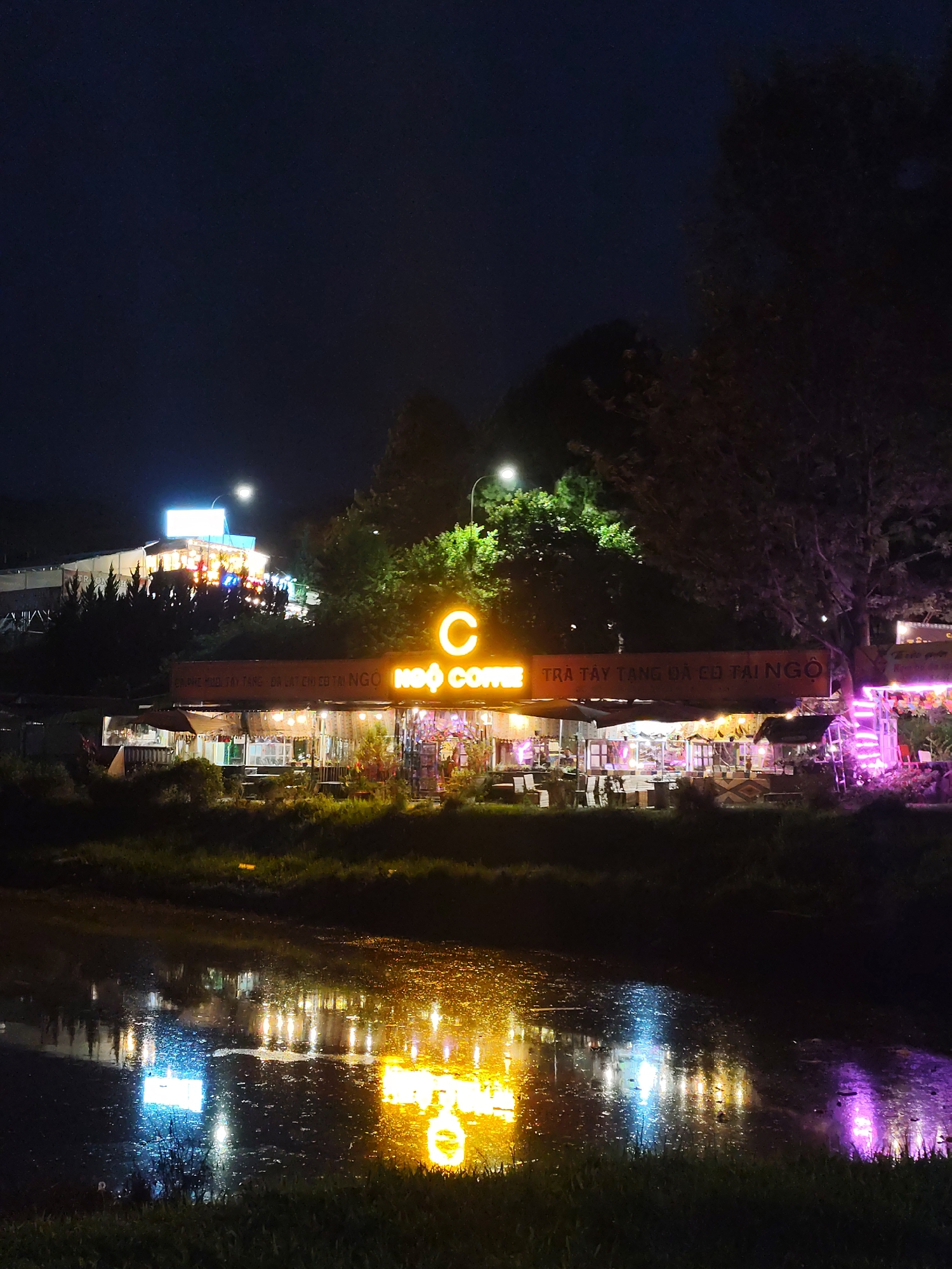 The image shows a night scene in Da Lat, Vietnam, featuring the Ngo Coffee cafe. The cafe has bright yellow neon signs. The cafe is reflected in the water in the foreground. The water also reflects the bright lights from a nearby building. A street light is visible behind the cafe.