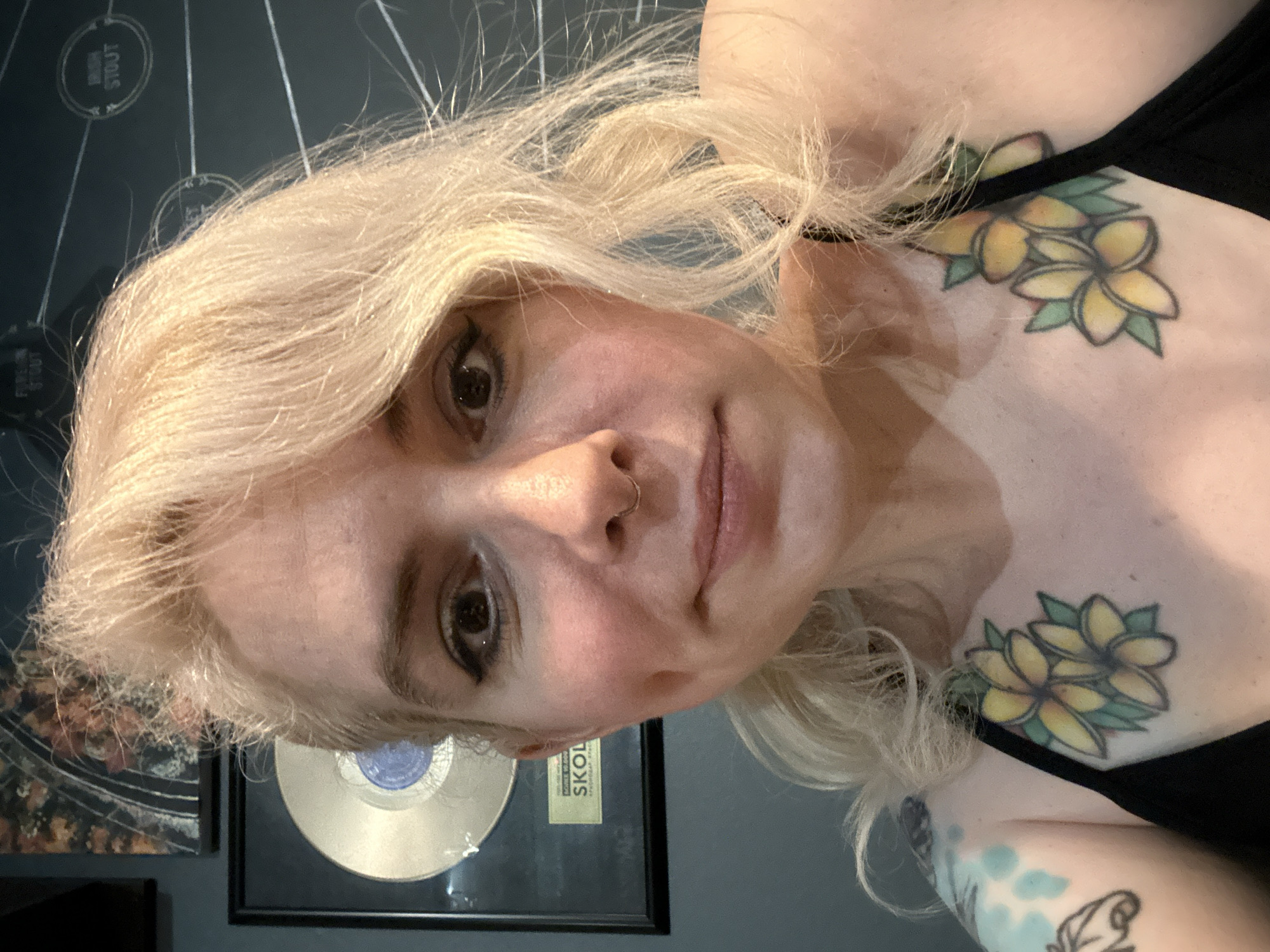 The image is a close-up, headshot of a young woman with blonde hair. She has a nose ring and is wearing a black top. She has tattoos of yellow flowers on her chest. In the background, there are framed items on the wall, including what appears to be a gold record and a framed poster or artwork. The overall lighting suggests an indoor setting, possibly during the day or evening. The woman's expression is neutral. Text is visible on the framed items in the background, including the words "IRON STOUT" and "SKOL" on a rectangular plaque.