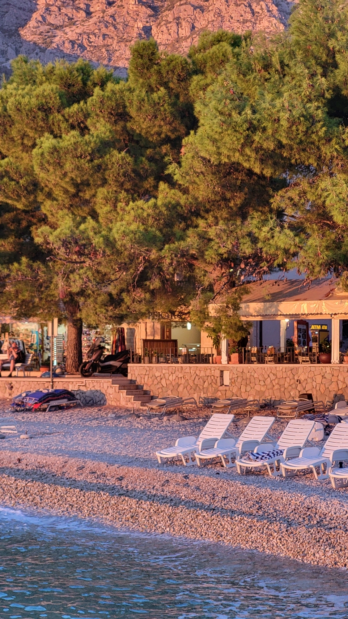 The image depicts a serene beach scene in Općina Tučepi, Croatia, bathed in the warm glow of what appears to be sunset. In the foreground, calm, turquoise water laps gently onto a pebble-strewn beach. A row of white plastic lounge chairs, some with towels draped over them, are neatly arranged on the shore, inviting relaxation. Further back, a stone-built restaurant or cafe with a light-colored awning overlooks the beach. Behind the restaurant, lush green pine trees dominate the mid-ground, their branches reaching towards a rugged, pink-hued mountain in the background. A single black scooter is parked near a tree on the beach, and several folded loungers and a blue inflatable object are stacked nearby. One person is visible sitting on a chair outside the establishment, while others are likely inside or out of frame, obscured by the trees and architecture. The overall atmosphere is one of tranquility and natural beauty.