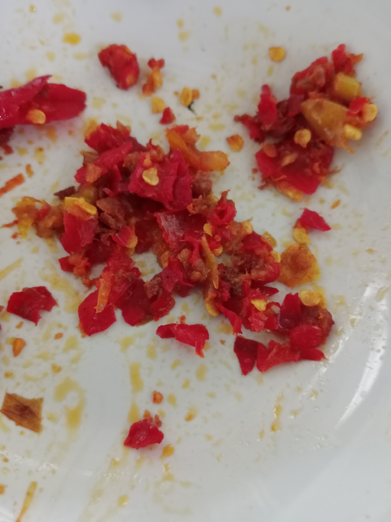 The image displays a close-up, overhead view of a white plate with remnants of chopped red chili peppers and their seeds scattered across it. The chili pieces are small and irregular in shape, with some appearing slightly wilted. Yellowish liquid, likely oil or sauce, is visible in streaks and pools on the plate, indicating that the chilies were part of a dish. The overall impression is that of a used plate after a meal. The scene is not indicative of any specific location in West Jakarta, Indonesia, as it lacks any recognizable landmarks or context.