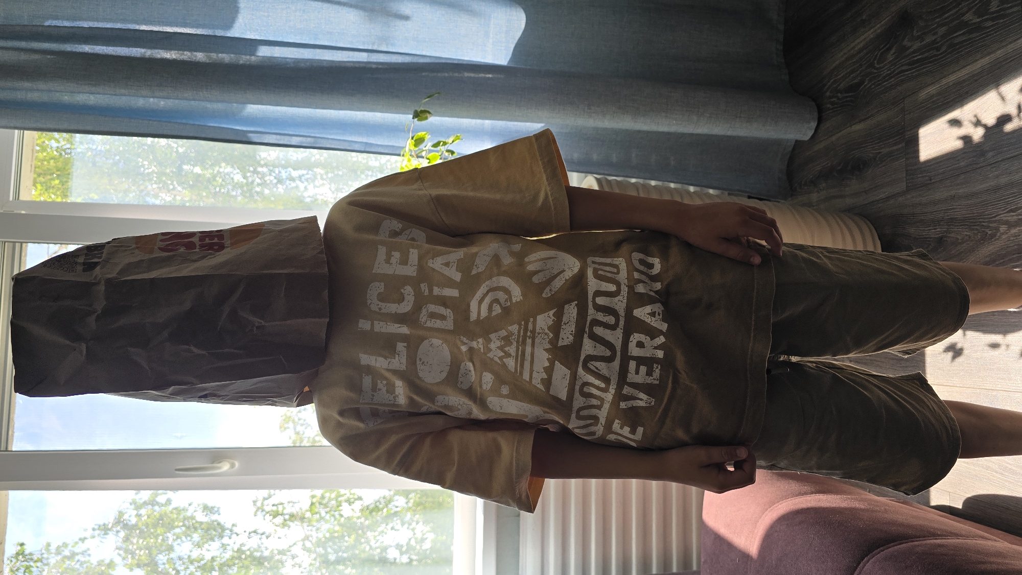 An individual, appearing to be a child, stands centrally in a well-lit indoor space within the city of Oktyabr'skii, Russia. The person's face is completely obscured by a crumpled brown paper bag, likely from a Burger King restaurant, with remnants of the fast-food chain's logo and branding visible on its surface. The individual wears a light tan or yellowish-brown short-sleeved t-shirt with distressed white text and graphics, reading "FELICES DÍA" (Happy Day) at the top and "DE VERANO" (of Summer) at the bottom, interspersed with abstract tribal-style patterns, including a stylized teepee or mountain, circular motifs, and handprint-like shapes. Below the t-shirt, the person wears dark olive green or brown shorts. The hands are visible at the sides, hanging naturally.
The background features a large window on the left, admitting significant natural light, which highlights green foliage and parts of trees outside. A clear sky is visible through the upper portion of the window. On the right side of the frame, a translucent light blue or grey-blue curtain is partially drawn, partially obscuring a view that includes more greenery and a light-colored, possibly woven, cylindrical object on the floor. A portion of a pink or mauve upholstered armchair or sofa is visible in the bottom-left corner. The floor is covered with dark wood-laminate or engineered wood, onto which distinct shadows are cast from the window frames and what appear to be leaves, indicating strong direct sunlight.