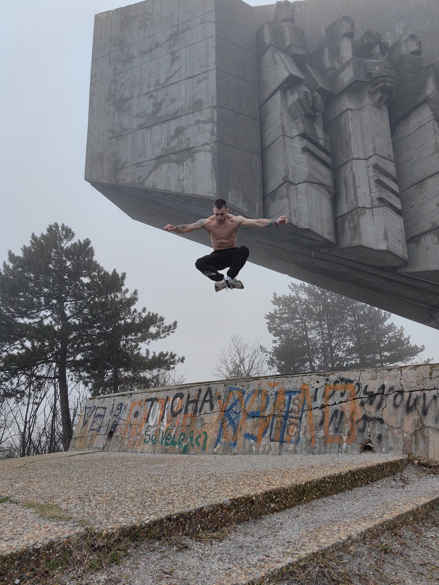 A shirtless man jumps in front of the Monument of the Bulgarian-Soviet Friendship in Varna, Bulgaria. He wears dark athletic pants and light-colored athletic shoes. His arms are extended to the sides. He jumps above a concrete wall covered in graffiti. Behind him, the monument is partially obscured by fog. Two pine trees stand to the left. The foreground shows a gravelly surface with two concrete steps.