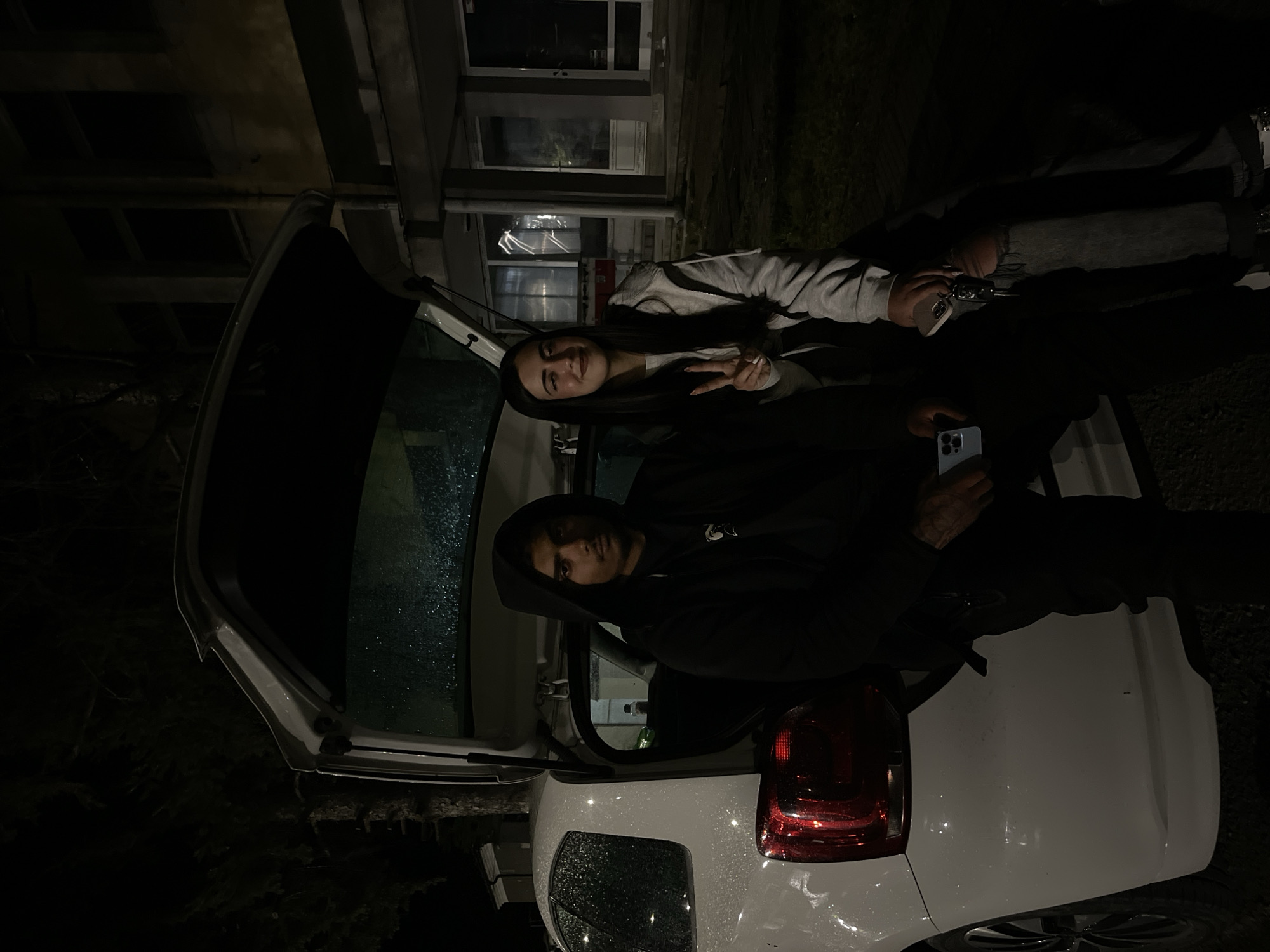 At night in Lozarevo, Bulgaria, a white car's open trunk is the focal point. Two individuals are near the car. A woman is making a peace sign, while a man in a hoodie holds a cell phone. The scene includes buildings in the background, visible through the car's open trunk. The area appears dimly lit, with rain droplets visible on the car's surface.