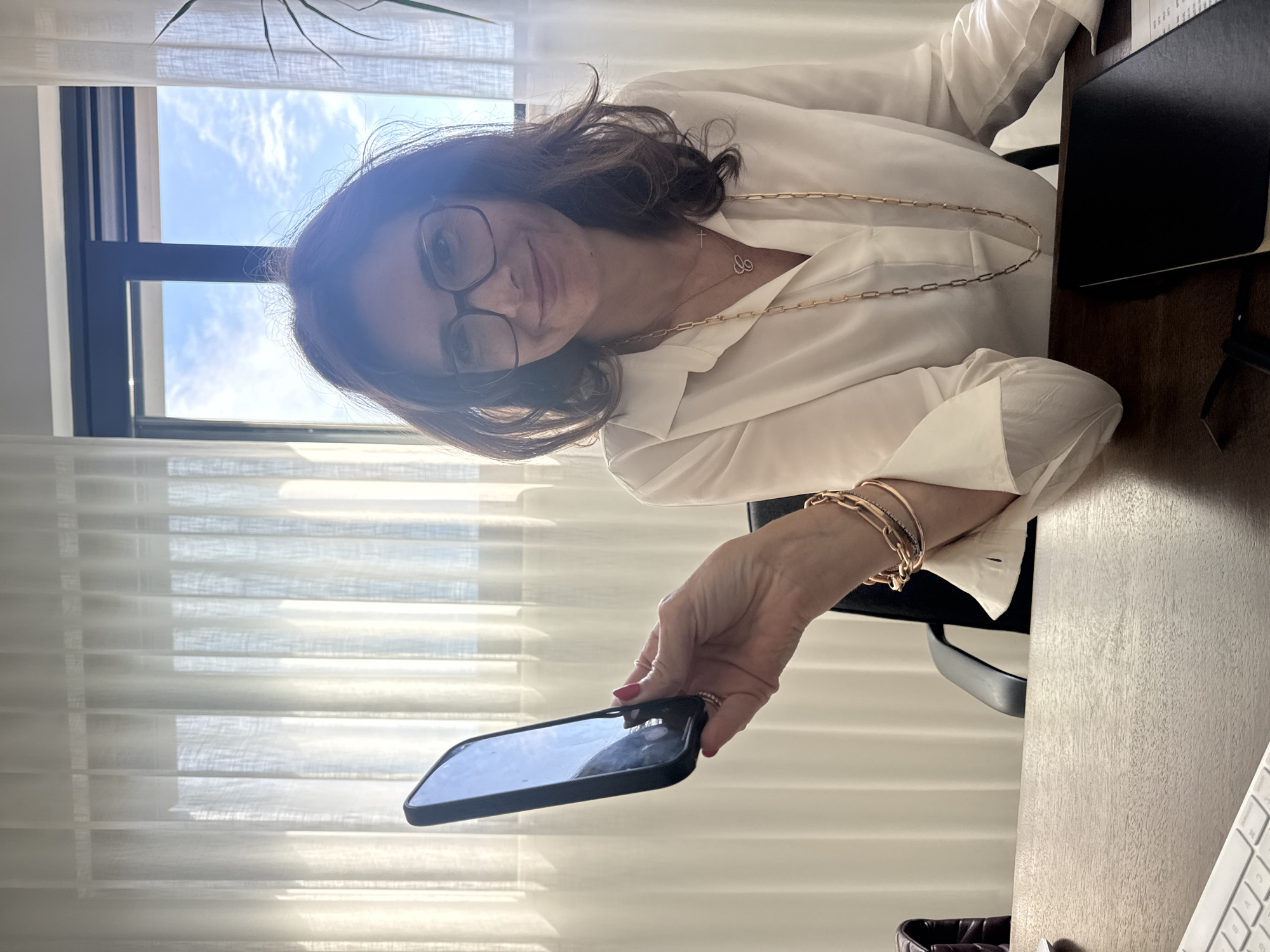 A woman wearing glasses and a white blouse is holding a smartphone. She is wearing gold bracelets and a gold necklace. She is seated at a desk with a notebook and keyboard visible. A window with blinds is behind her. The location in Torres Vedras, Portugal cannot be determined from the image.