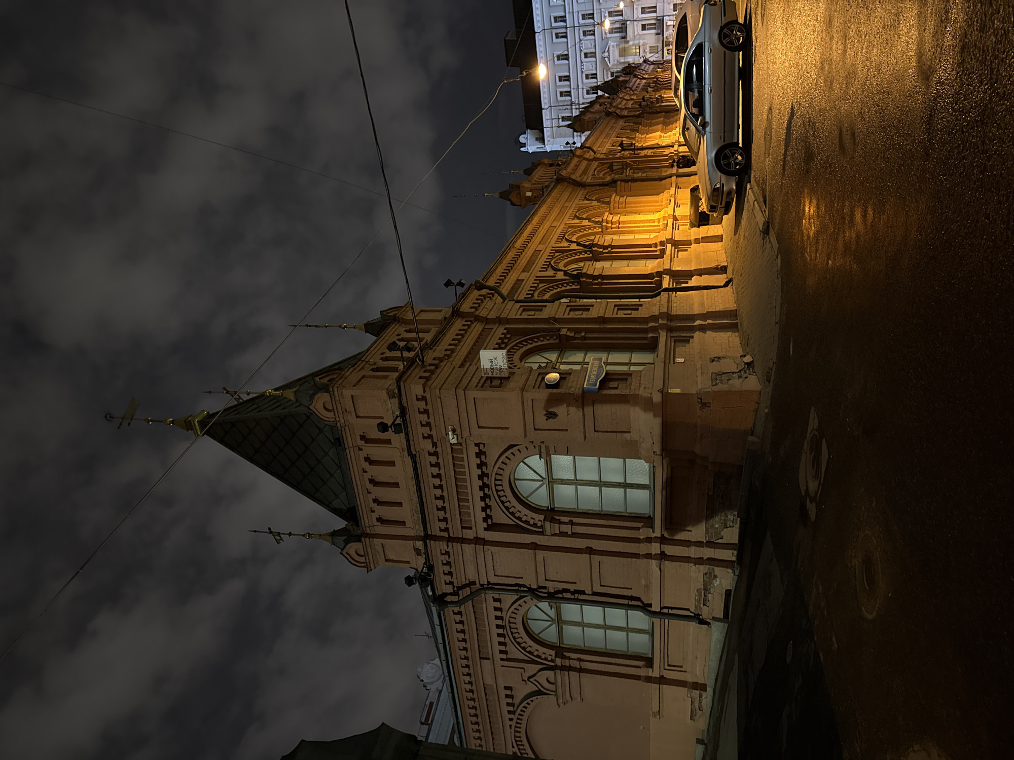 The image shows a view of the Perlov Tea House in Moscow, Russia at night. The building is ornate, with decorative brickwork, arched windows, and a distinctive pagoda-style roof. A parked car is on the right side of the frame, illuminated by streetlights, which reflect off the wet pavement. The sky is cloudy and dark. There are power lines visible in the upper portion of the frame.