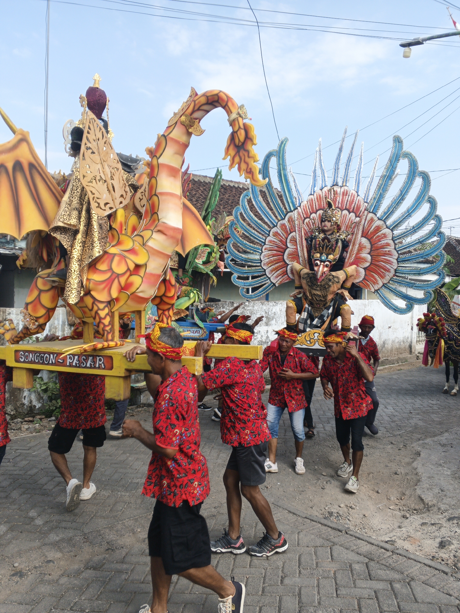 A procession of people in Singolatren, Indonesia, carries large, elaborate floats. The procession moves along a paved street lined with buildings and vegetation. The sky above is blue with some clouds. In the foreground, several men wearing red shirts with a black floral pattern and headbands are carrying a yellow float with the inscription "SONGGON PASAR". This float supports a large, ornate dragon figure. Further back, another float features a figure resembling a bird or mythical creature with a large, colorful headdress and blue, red, and white feathered wings, also carried by men. A horse decorated with elaborate tack and colorful tassels is visible on the right side of the image. The overall atmosphere suggests a festive or cultural event.