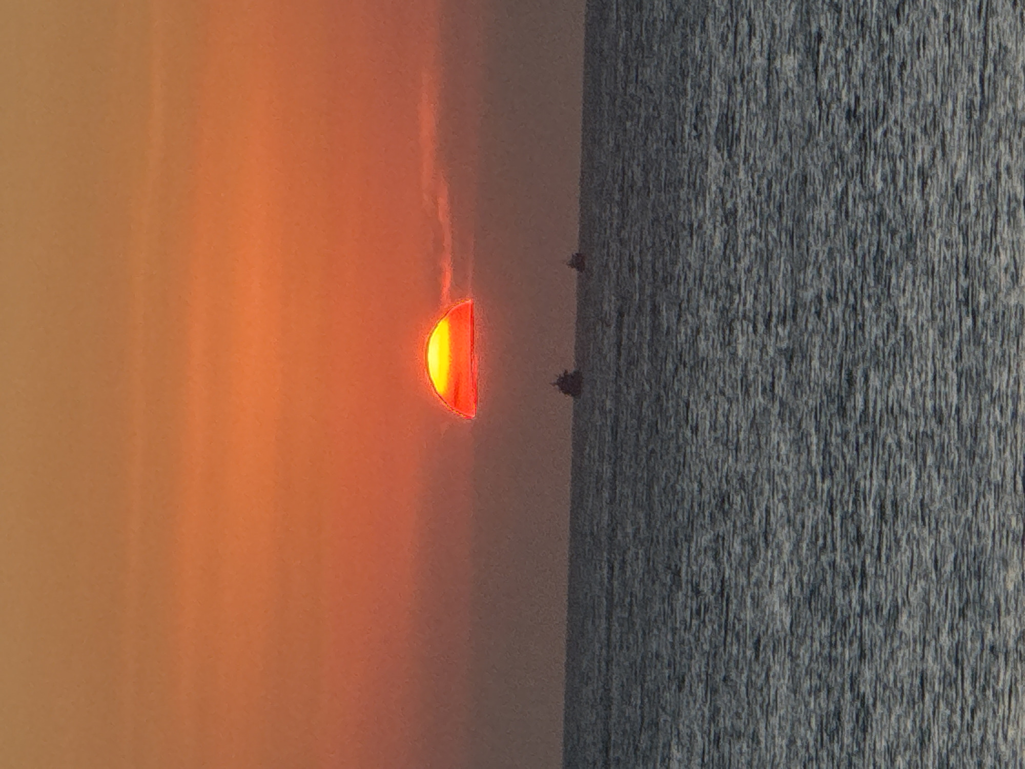 This image captures a vibrant sunset or sunrise over the ocean, consistent with the coastal setting of Kuta Selatan, Indonesia. The scene is dominated by a large, intensely bright, semi-circular sun positioned on the horizon, partially submerged beneath the water's surface.
The sky is filled with a dramatic gradient of warm colors, transitioning from brilliant yellow and orange immediately around the sun to richer reds and deeper oranges towards the top of the frame. Faint, wispy streaks of red light extend upwards from the sun, suggesting atmospheric haze or clouds catching the light.
Below the distinct horizon line, the ocean fills the lower part of the image. The water surface appears dark with a textured, slightly choppy quality, characterized by numerous small, vertical ripples or reflections. Two small, dark silhouettes, possibly distant fishing boats or very small land formations, are visible on the horizon, silhouetted against the bright glow of the sun. The overall scene is one of tranquil natural beauty, highlighting the striking colors and vastness of the sea at either dawn or dusk. There is no visible text in the image.
