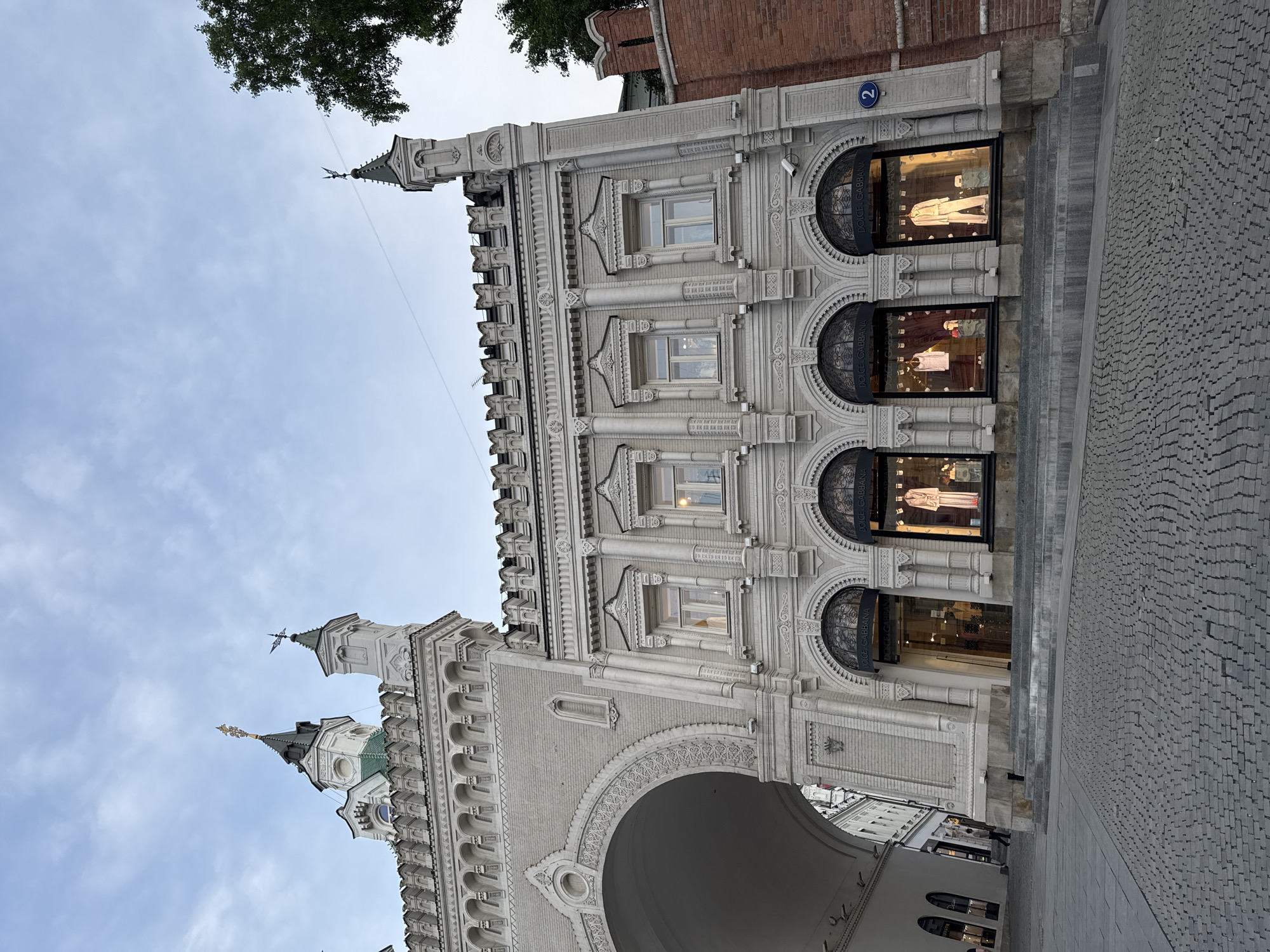 The image captures a detailed view of the Resurrection Gate in Moscow, Russia, particularly emphasizing the facade of the adjacent building which houses a Dolce & Gabbana store. The architecture is elaborate with intricate ornamentation and arched windows.
The building facade is made of light-colored brick and includes three levels of windows, with each level featuring decorative detailing. A small street sign labeled "2" can be seen near the top right corner of the building facade. Below the windows are awnings with "DOLCE & GABBANA" printed on them.
The foreground shows the paved walkway leading through the arched entrance of the Resurrection Gate. The sky above is partly cloudy.