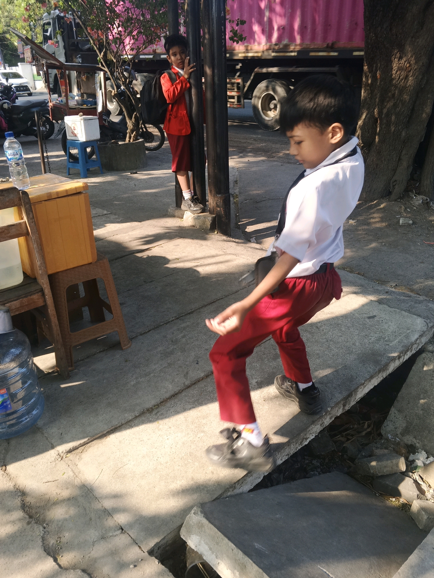 In Krian, Indonesia, two young boys are captured in a sun-drenched outdoor scene. The boy in the foreground, wearing a white school shirt and red pants, is in mid-motion, his body angled as if dancing or posing. His right leg is extended forward, and his arms are bent at the elbows, with his right hand slightly raised. He is wearing dark sneakers and white socks, with a dark bag slung across his shoulder. He is standing on a concrete surface that appears to be a raised walkway or sidewalk, with a visible gap or trench next to it.
Behind him, partially obscured by a tree and a dark pole, stands another boy. He is wearing a red school jacket over a white shirt and a red skirt or shorts. He has a black backpack on. He is also wearing dark shoes and white socks. He is looking towards the camera and has his right hand raised in a gesture, possibly a peace sign or a wave.
The environment suggests a public space in Krian, with elements of urban life. In the background, a large truck with a pink cargo container is visible, as well as other parked motorcycles. A street vendor's cart with a yellow box and a cooler sits to the left of the foreground boy, with a large blue water jug next to it. The ground is a mix of concrete and dirt, with trees and shadows casting patterns from the sunlight. The overall impression is a candid snapshot of children in a bustling, everyday setting.