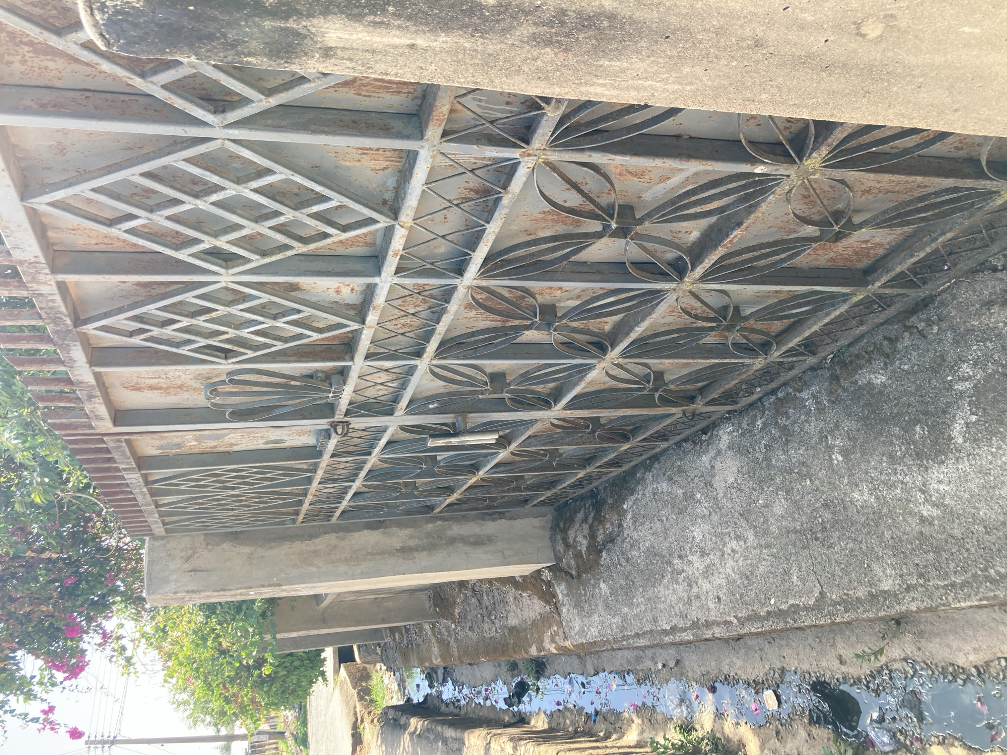 The image depicts a wrought iron pedestrian bridge in Jos, Nigeria. The bridge spans a drainage ditch filled with stagnant water and debris. The ironwork shows signs of rust. The bridge is supported by weathered concrete abutments. Vegetation is visible near one end of the bridge. The environment is urban, and the presence of stagnant water indicates potential sanitation issues. No people are visible.