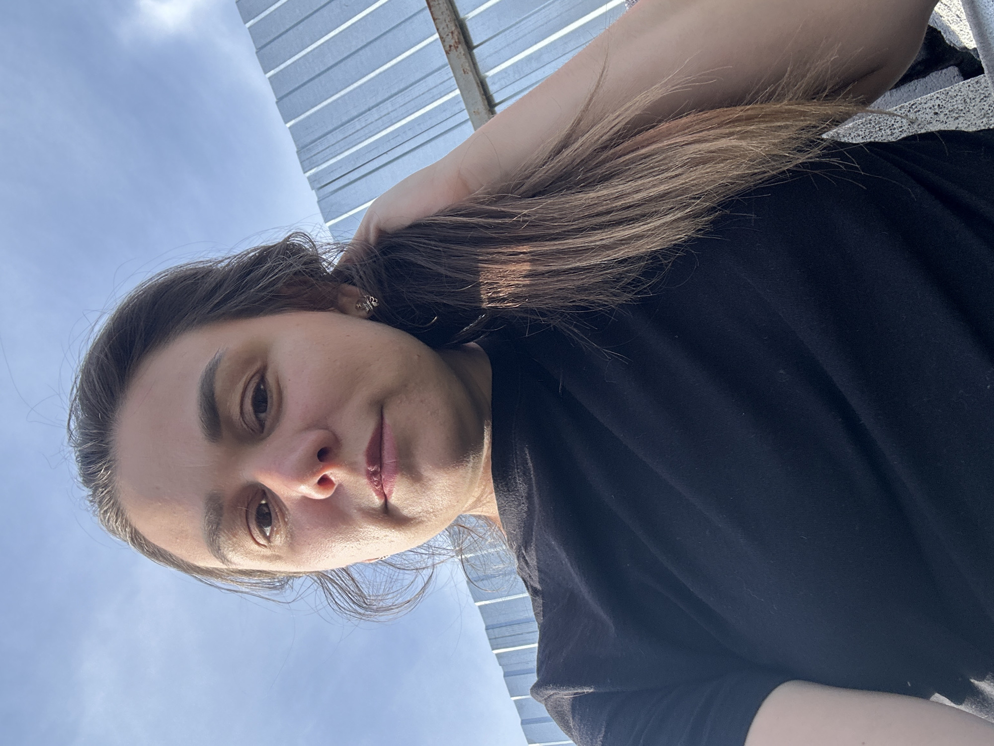 This is a selfie taken by a person outdoors, likely on a sunny day, given the bright sky and shadows. The camera is held above the person's head, pointing downwards, creating a unique perspective. The person, whose face is partially visible, has long, dark brown hair that is being held up with their right hand. They are wearing a black t-shirt.
In the background, there are two distinct elements. To the left, a clear blue sky with some wispy white clouds is visible, suggesting fair weather. To the right, a structure made of corrugated metal, possibly a roof or wall, is seen with distinct horizontal lines. This structure appears to be on top of what looks like a concrete surface, possibly a rooftop or a balcony.
The lighting suggests it's daytime, with sunlight casting shadows and illuminating the scene. The person's expression is neutral, but the upward tilt of the head and the way they are holding their hair might imply a casual or relaxed pose. The image doesn't contain any visible text. The overall impression is a candid shot taken in an outdoor urban or semi-urban environment.