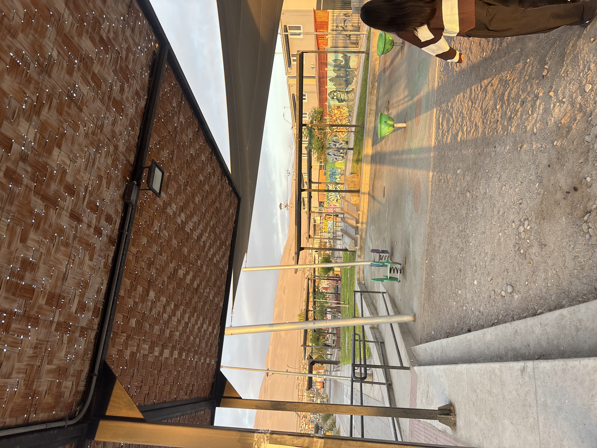 The image captures a view from a shaded structure, possibly a pergola, towards a children's playground in the city of Arica, Chile. The playground features several elements, including green seesaws, spring riders, and graffiti-covered walls along a building in the background. In the distance, a large sandy hill or dune is visible against a cloudy sky. The foreground shows concrete steps leading down from the structure, and a person with dark hair and a striped sweater can be seen at the edge of the playground. The time of day appears to be late afternoon or early evening, based on the warm light and long shadows cast across the scene.