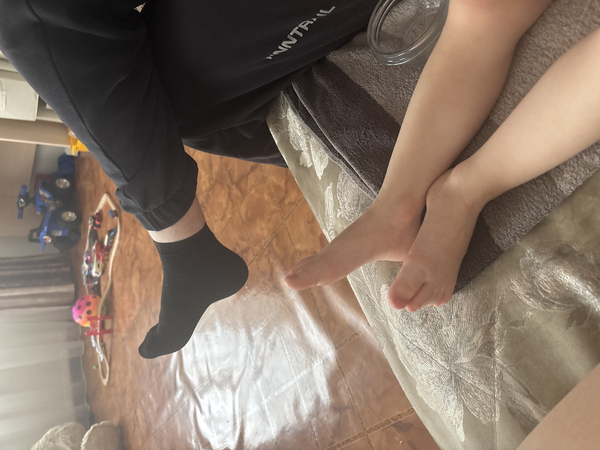 This media file captures a close-up, low-angle view of two individuals' legs and feet in what appears to be an indoor home setting, possibly in Ferma, Russia.
**Subjects:** Two people are visible: an adult and a child. The adult, positioned on the left, is wearing black sweatpants with partially visible white text that reads "WNNTRAIL" or similar, and a black sock on their visible foot. The child, on the right, has bare, light-skinned legs and feet, which appear to be resting or crossed on a cushioned surface.
**Objects:** The adult's black sweatpants are a prominent item of clothing. A black sock is visible on one of the adult's feet. The child's bare skin is notable. Both individuals are partially on or next to a piece of furniture covered by a grey and off-white patterned blanket or cushion, featuring a floral design. A clear, round glass object, resembling a bowl or cup, is visible near the child's legs on this surface. The floor beneath is covered with light brown or tan square tiles. Scattered on the tiled floor are children's toys, including a blue ride-on toy truck and a small pink and yellow ball. A light-colored curtain or sheer fabric is partially visible on the far left, suggesting a window.
**Activity or Event:** The individuals appear to be relaxed, possibly sitting or reclining on a couch or bed. There is no specific activity evident other than resting.
**Notable Details:** The image is well-lit by natural light, indicating it is daytime. No specific weather conditions are discernible. The setting appears to be a casual living space or bedroom. The text "WNNTRAIL" on the adult's pants is a visible detail. The general atmosphere is calm and domestic.