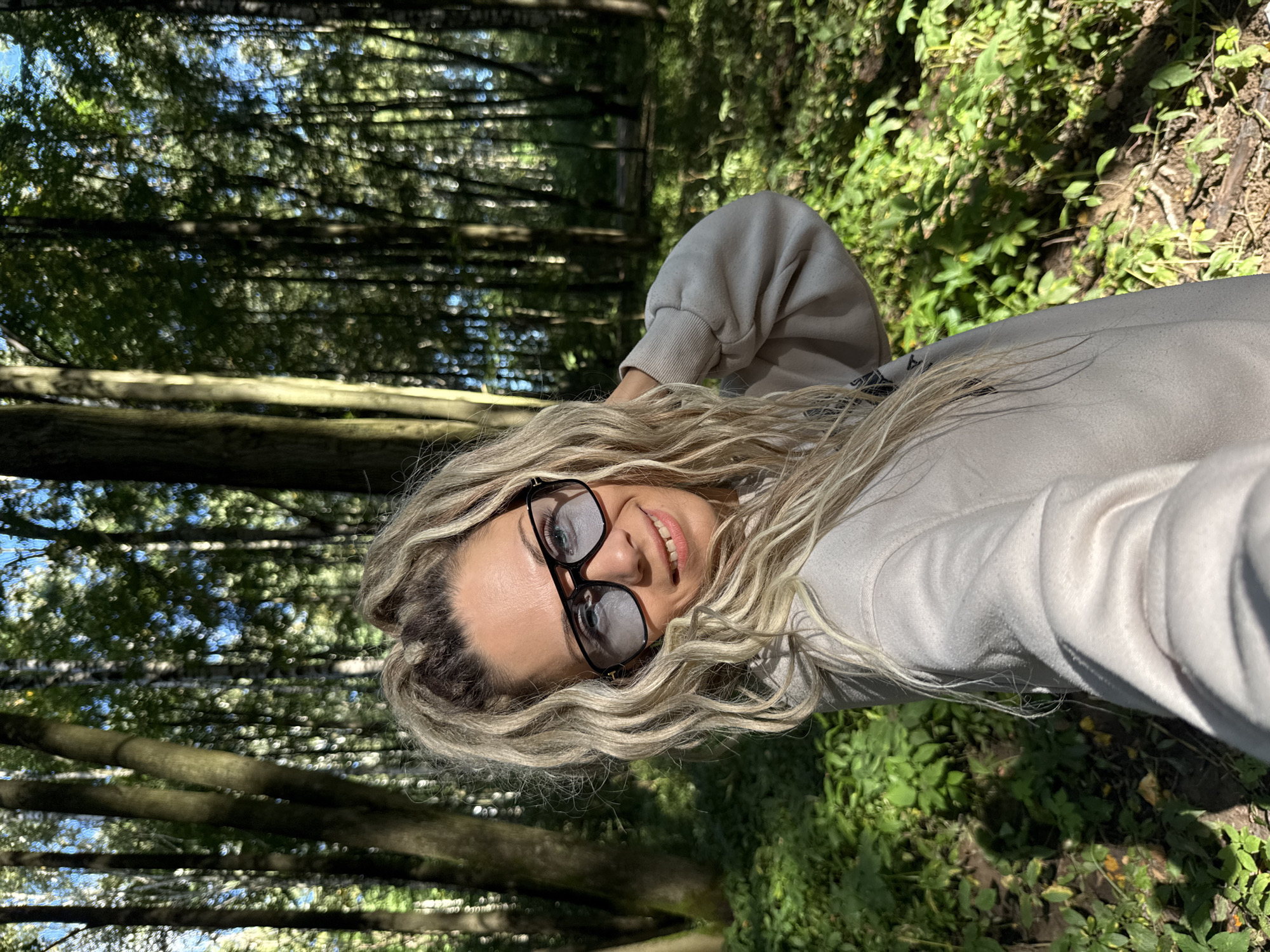 A selfie is taken by a woman in a light-colored hooded sweatshirt in a forest. She has long, wavy blonde hair and is wearing large, dark-rimmed glasses with light blue lenses. Her right hand is touching her head, and she is smiling at the camera. The background is a dense forest with tall trees and green undergrowth, dappled with sunlight. The image appears to have been taken outdoors in a natural setting, possibly in the vicinity of Rusanovo, Russia, given the type of deciduous forest typical of the region. The lighting suggests it is daytime, with the sun filtering through the trees.