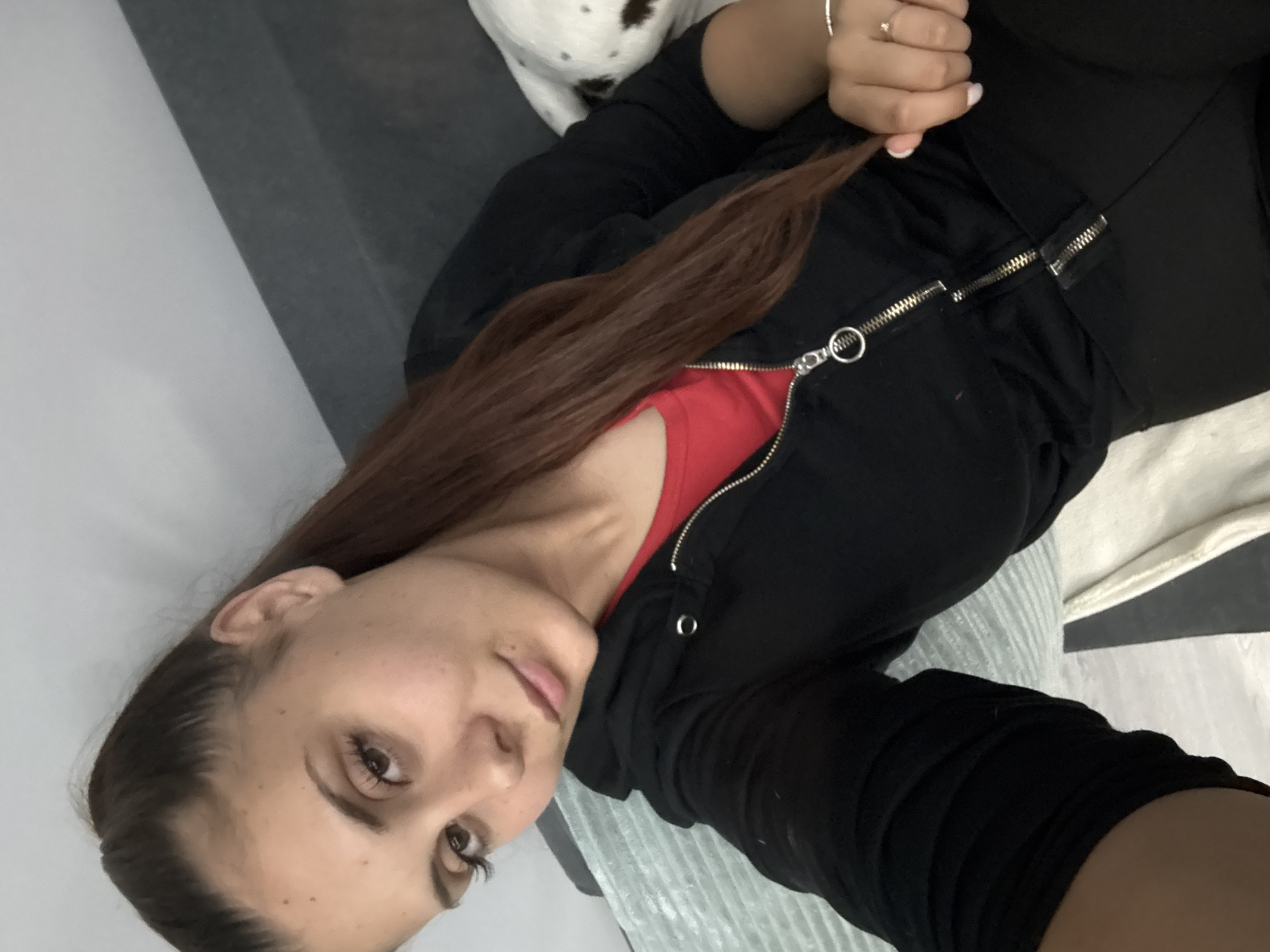 A young woman is depicted lying down, angled towards the camera, likely taking a selfie, in an indoor setting in Elin Pelin, Bulgaria. Her head is tilted, and she looks directly at the camera with a calm, relaxed expression, possibly with a subtle smile. She has long, straight, reddish-brown hair that falls across her left shoulder and chest. Her eyes are dark, and she wears natural-looking makeup.
She is dressed in a black zip-up hoodie, worn open to reveal a bright red top underneath. On her left hand, which is resting near her chest and holding a strand of her hair, she wears a ring with a light-colored stone and possibly a simple band. Her fingernails feature a light-colored polish.
In the upper left background, partially visible behind her shoulder, is a white animal with black spots, strongly suggesting a dog like a Dalmatian. The immediate setting appears to be a bed or couch, characterized by neutral-colored fabrics in white and gray tones. The lighting is soft and even, indicating an indoor environment, either during the day or in a well-lit room. There is no visible text in the image.