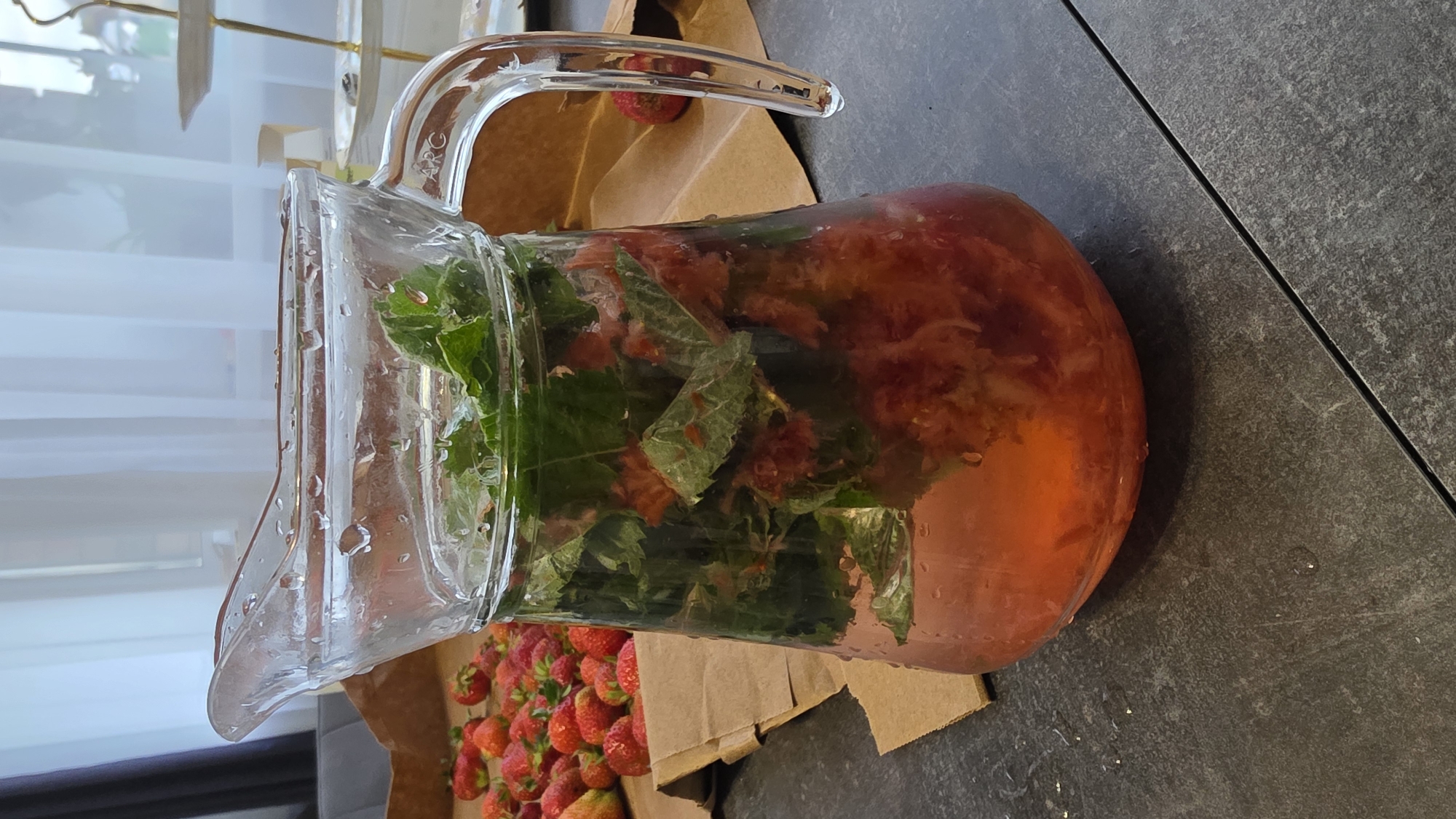 A clear glass pitcher filled with a pink-hued liquid containing chopped strawberries and mint leaves sits on a dark gray textured surface. Numerous fresh strawberries are scattered on brown paper behind the pitcher. The background shows a dimly lit room with sheer white curtains and a glimpse of furniture. The scene is likely indoors, given the interior setting. The location is identified as Bulgakovo, Russia.