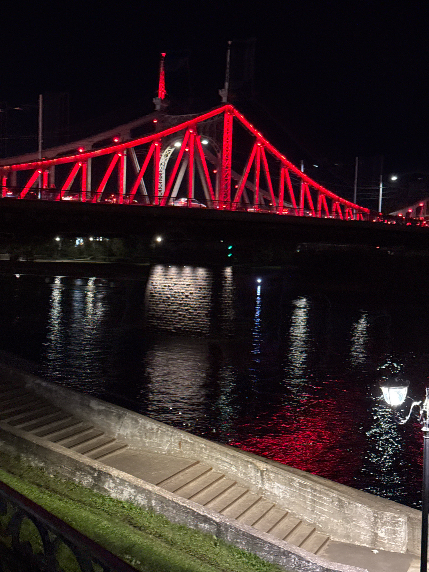 This is a nighttime photograph of the Starovolzhsky Bridge in Tver, Russia, illuminated with red lights. The bridge is a prominent feature, with its steel structure visible and outlined in bright red. Cars are moving across the bridge, indicated by their headlights and taillights. Below the bridge, a body of water, likely the Volga River, reflects the bridge's lights, creating shimmering patterns on the dark surface. On the left side of the foreground, there are stone steps leading down to the water's edge, bordered by a grassy area and a dark railing. A vintage-style lamppost stands on the right side of the foreground, its light illuminating the immediate area. The overall scene depicts a well-lit architectural landmark at night, with its reflection on the water adding to the visual appeal.