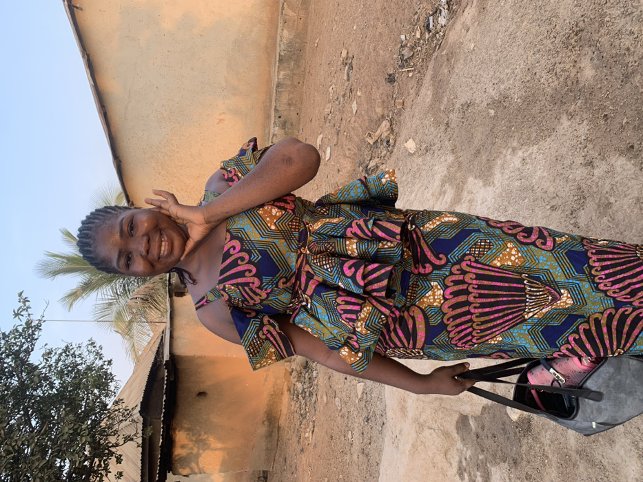 A smiling woman in a vibrant, patterned dress poses outside. The dress features a mix of navy, teal, brown, and pink designs, cinched with a matching sash. She holds a bag, and a small house with a corrugated metal roof is partially visible in the background, along with some vegetation. The terrain appears to be dirt and dust. The sky is bright and clear.