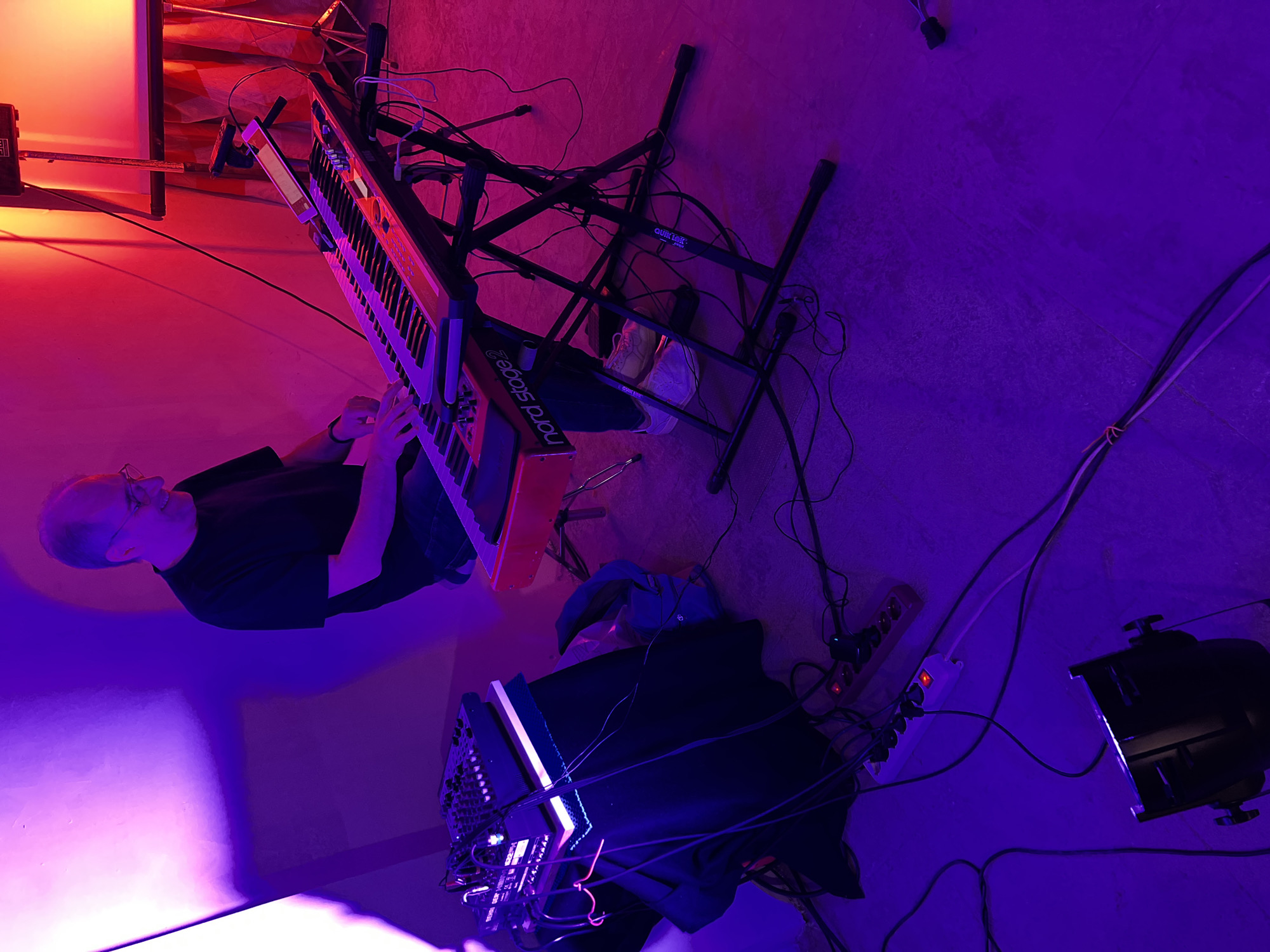 This image captures an indoor musical setup in Santeramo in Colle, Italy, illuminated by strong colored lighting. A middle-aged male, wearing a black t-shirt and glasses, is seated on a stool, actively playing a red Nord Stage 2 keyboard synthesizer positioned on a black X-style stand. He is smiling and looking down at the keys, using both hands. To his left, a small black Behringer audio mixer, connected with multiple cables, rests on a stack of white objects covered by dark fabric. Numerous black and white audio and power cables are visible strewn across the concrete floor, connecting various pieces of equipment, including two multi-outlet power strips, one white and one brown, both showing active red indicator lights. A black stage light fixture is present on the floor in the lower left foreground. In the background, on the right, a white projection screen is visible, with red fabric or drapes behind it, and a speaker or monitor is mounted on a stand. The scene is bathed in vibrant purple light on the left side, transitioning to a red hue on the right, indicating a dynamic stage or studio lighting arrangement.