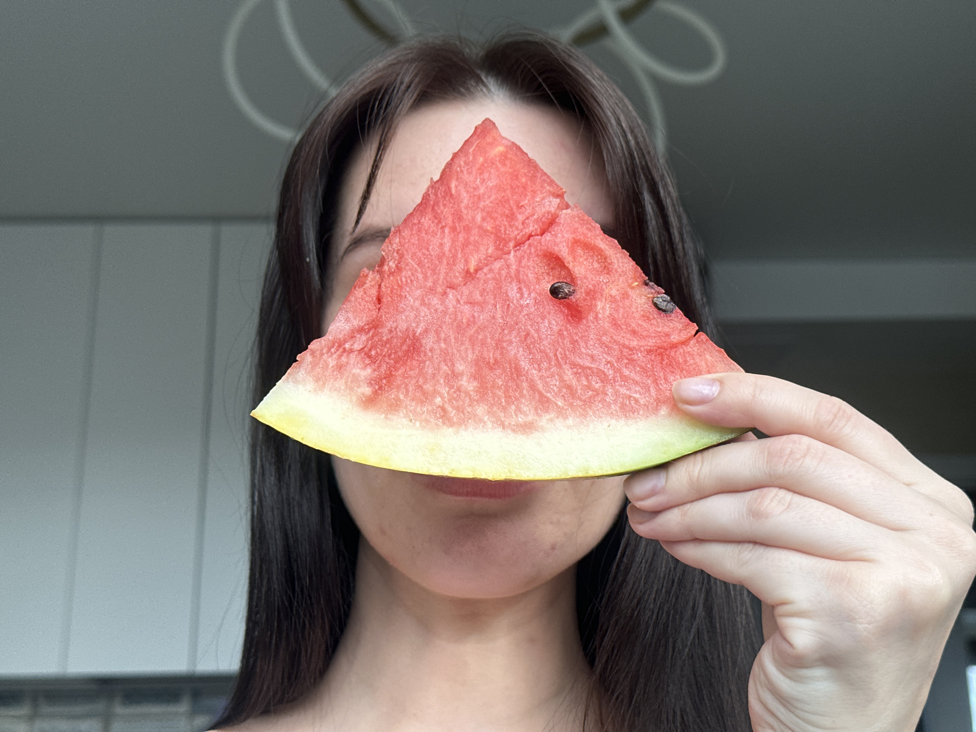 A woman holds a slice of watermelon in front of her face indoors. The watermelon is a triangular slice with red flesh and black seeds. The woman has long, dark hair. The background shows light-colored cabinets and a chandelier. The photograph was likely taken in Chaikovsky, Russia.
