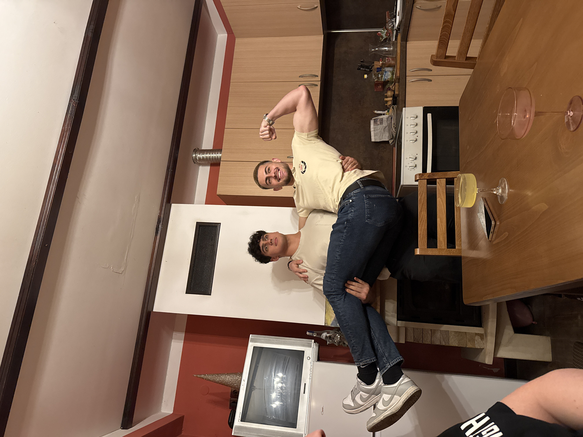 Two men are in a residential kitchen. One man is lifting the other; the lifted man is flexing his bicep. The kitchen has wooden cabinets, a white oven, and a wooden table with glasses. The environment includes a fireplace, a television set, and exposed ceiling beams. No information in the image indicates this location is Berkovitsa, Bulgaria.