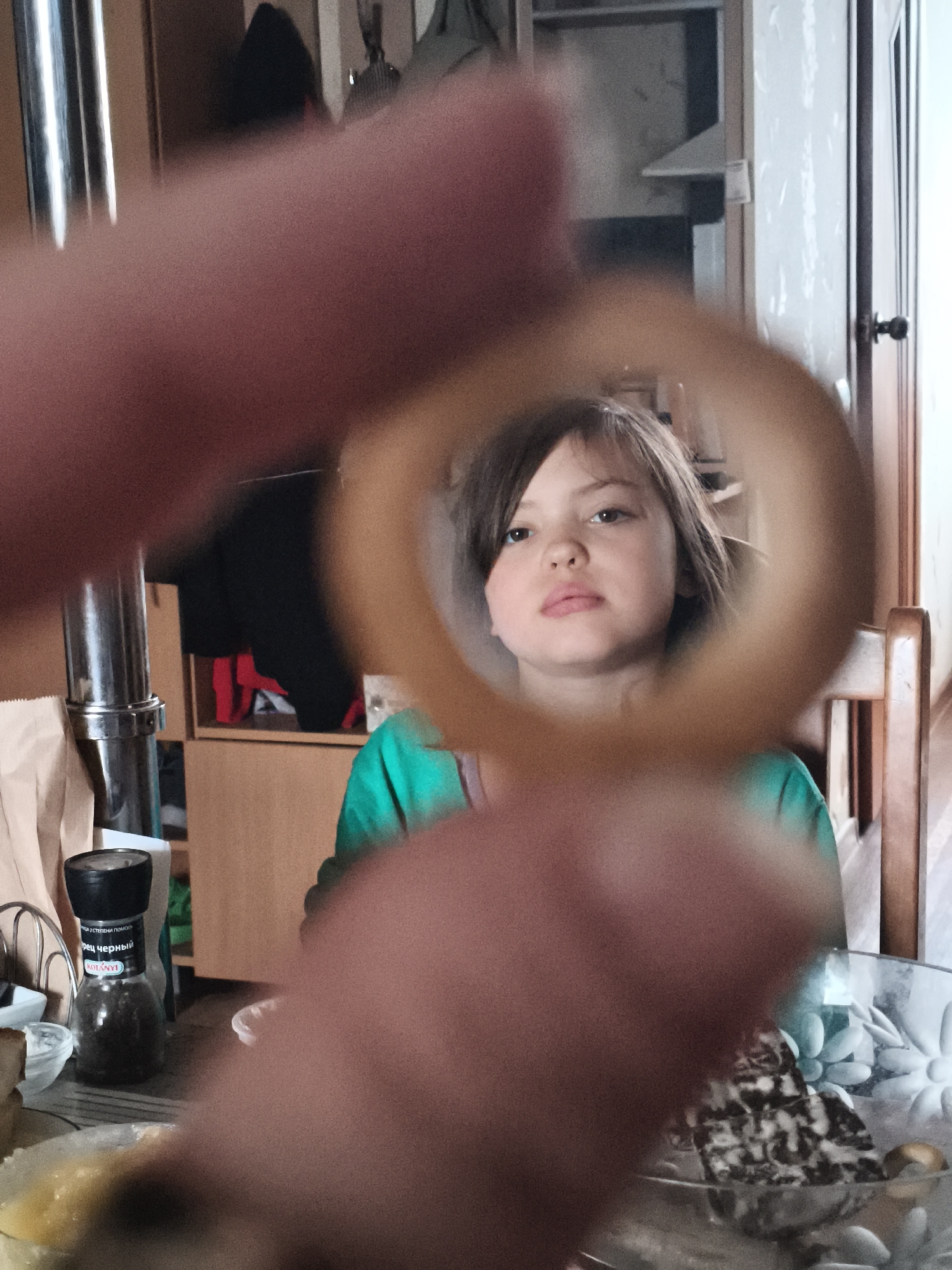 The image presents a close-up, first-person perspective photograph captured indoors, presumably in a domestic setting like a dining area in the city of Unknown, Unknown. The foreground is dominated by a blurred human hand, depicted as reddish-pink flesh, holding a light brown, circular object, possibly a food item such as a pretzel ring, directly in front of the camera lens.
Through the opening of this ring, the face of a young Caucasian female child is framed in the midground. She has shoulder-length, dark brown hair and a neutral expression with slightly parted, pink lips. Her dark-colored eyes are directed upwards and slightly to the viewer's left. She is wearing a teal-colored top.
The background shows various household elements. To the immediate left of the child, a metallic cylindrical pipe runs vertically, partially obscured by a light brown paper bag. A black pepper grinder, labeled "KOTANYI" and "ПЕРЕЦ ЧЕРНЫЙ" (Black Pepper) in Cyrillic, rests on a surface, next to partially visible plates containing yellowish food. In the mid-background, wooden cabinetry or shelves display folded black and red garments. To the right of the child, a light-colored wooden chair back is visible, along with a white door featuring a dark doorknob. On a surface in the lower right, a clear glass bowl is filled with various light-colored, elongated confectionery items or candies, alongside some patterned, possibly baked, goods. The overall lighting is soft and ambient.