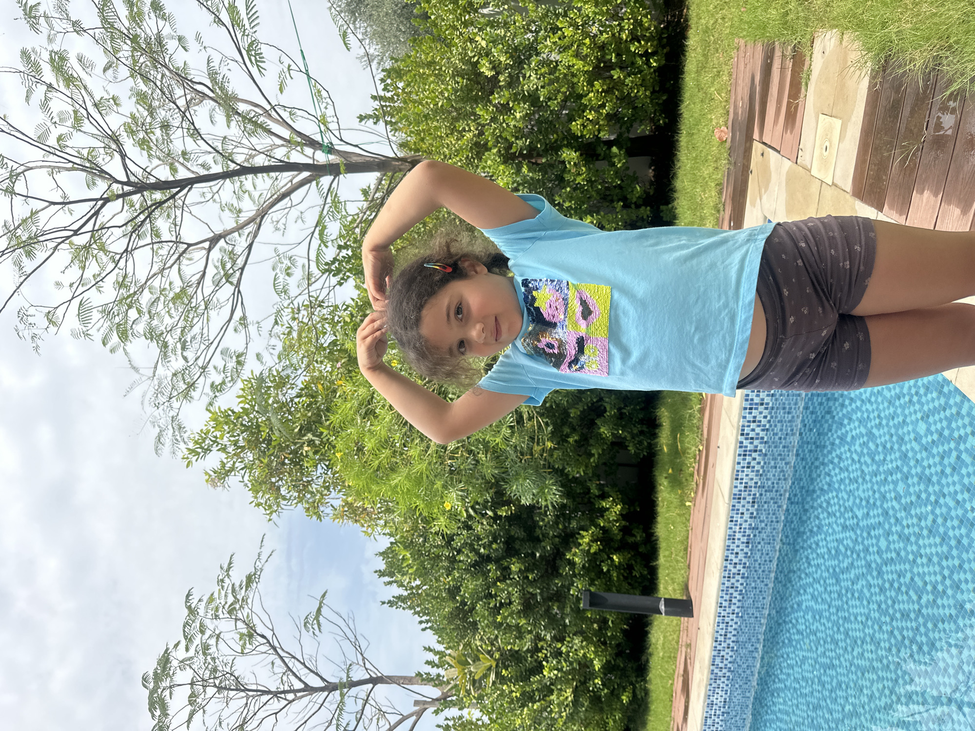 A young girl is captured in an outdoor setting, likely a private garden or backyard in Dubai, United Arab Emirates. The image, taken on a bright, partially cloudy day, shows the girl posing near a swimming pool.
The subject, a young girl with curly dark hair, looks directly at the camera with a gentle expression. She is dressed in a light blue short-sleeved t-shirt with a colorful sequined design on the chest, incorporating pink, yellow, blue, and black hues, possibly forming an abstract pattern or animal face. She pairs this with dark grey shorts adorned with a subtle light floral print. Her arms are raised and bent above her head in a relaxed pose, and a multi-colored hair clip is visible in her hair.
The background features a light blue tiled swimming pool, edged with light-colored paving and wooden decking. Lush green grass and dense foliage from various trees and bushes surround the area. A prominent tree with delicate, fern-like leaves stands against the bright sky, alongside another tree with broader, darker green leaves. A thin green rope or wire is strung between some branches. A dark, slender pole is also visible near the pool's edge. The overall scene suggests a pleasant and relaxed outdoor environment. There is no visible text in the image.