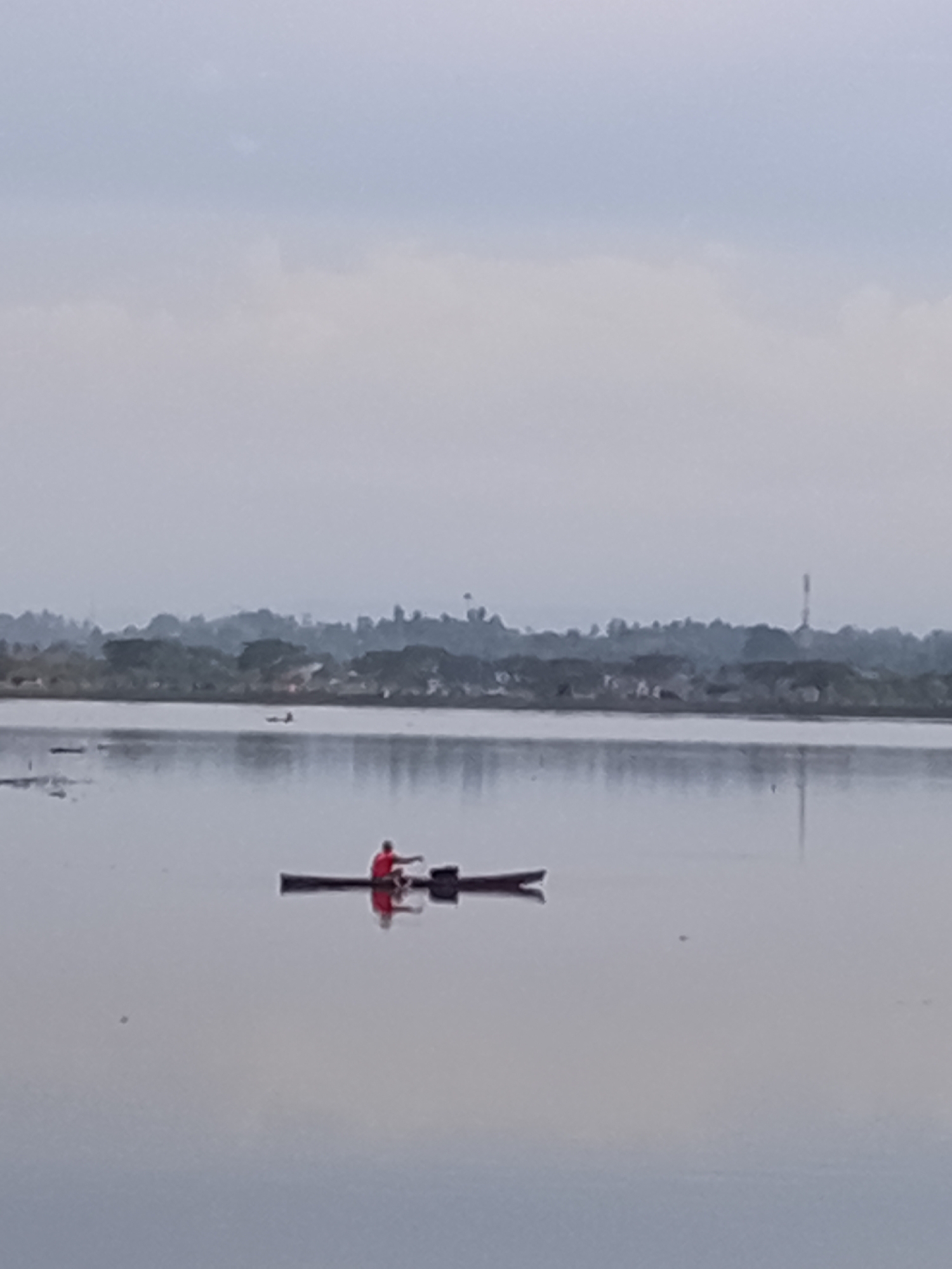 The image depicts a serene lake or calm body of water, likely Lhokseumawe, Indonesia. The sky is overcast and hazy, suggesting either early morning or late afternoon. The water's surface is still and reflective, mirroring the subdued tones of the sky and the distant landscape.
The primary subject is a person in a narrow, elongated boat, possibly a dugout canoe, on the water. The person is wearing a bright red shirt and appears to be engaged in paddling or steering the boat. The reflection of the boat and the person is clearly visible on the water's surface, creating a symmetrical, almost dreamlike effect. In the distance, a treeline with a scattering of buildings and a tall tower suggests a coastal or lakeside community. The overall mood is peaceful and tranquil, with a sense of solitude and quiet activity. The scene is set against a backdrop of soft, muted colors, emphasizing the vastness of the water and sky. There is no visible text in the image.